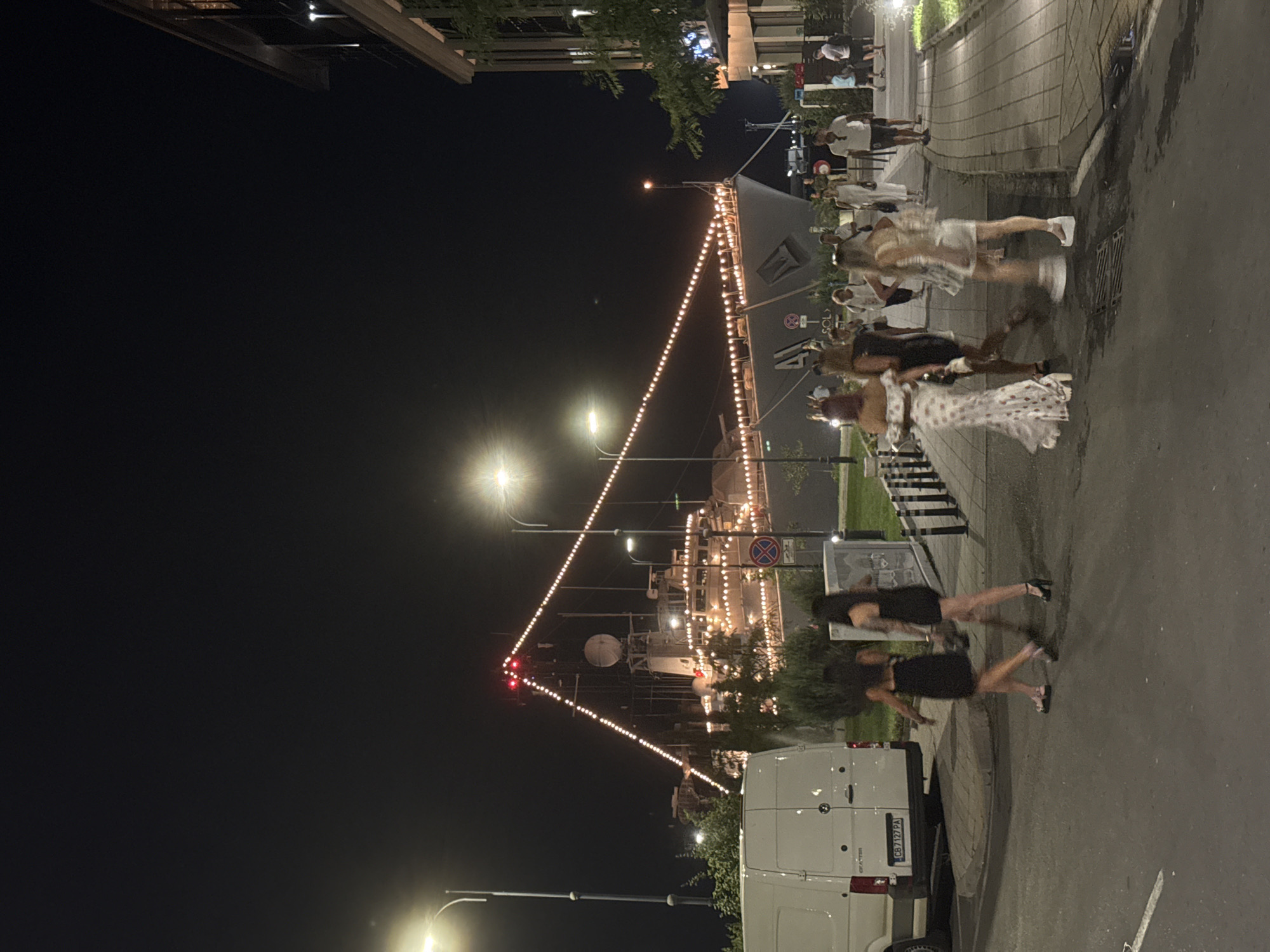 The image depicts the Burgas Sea Station in Burgas, Bulgaria at night. A large ship, illuminated by numerous string lights, is docked in the background. In the foreground, several people are walking along a paved pathway and a street. A white van is parked on the left side of the street, and a modern building with multiple levels is visible on the right side of the image. The overall atmosphere suggests a lively evening scene at a waterfront location.