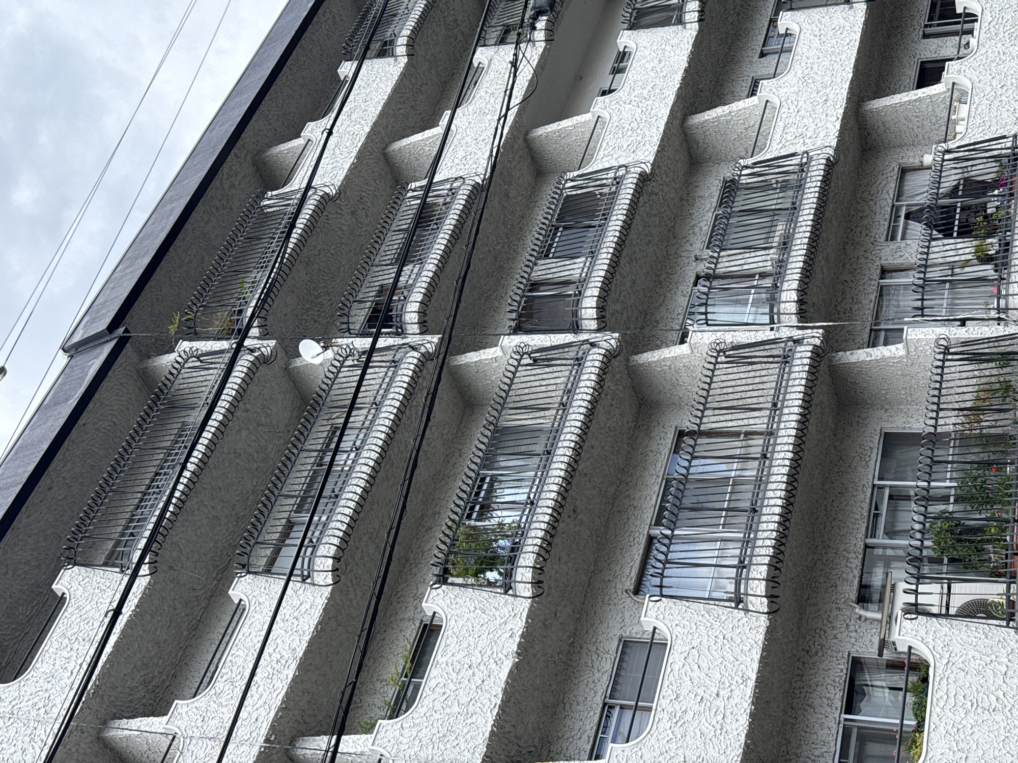 The image captures a worm's-eye view of a modern, multi-story building, likely an apartment or office complex, set against an overcast sky. The building's facade is textured with a light-colored, possibly white or pale grey, rough stucco finish.
A prominent and repetitive architectural feature consists of deep, recessed windows or balconies, each protected by an intricate, dark-colored metal grille. These grilles are composed of horizontal bars and distinct spiraling or coiled elements along their vertical edges, providing both a decorative and potentially protective function. The windows behind these grilles are large and reflective, showing glimpses of the cloudy sky and parts of the building itself. On some of the balconies, small potted plants are visible, adding touches of greenery to the urban structure. One balcony in the lower right features an outdoor air conditioning unit and another small plant, while a smaller, circular white satellite dish is mounted on a balcony higher up.
Multiple utility wires, dark in color, run diagonally across the foreground and midground, emphasizing an urban setting. The sky in the upper left corner is uniformly grey and cloudy, indicating an overcast day. No people are visible, and there is no discernible text on the building or in the surrounding environment. The architecture suggests a dense urban environment, typical of cityscapes such as Kyoto, with a focus on functional yet distinctive design elements.