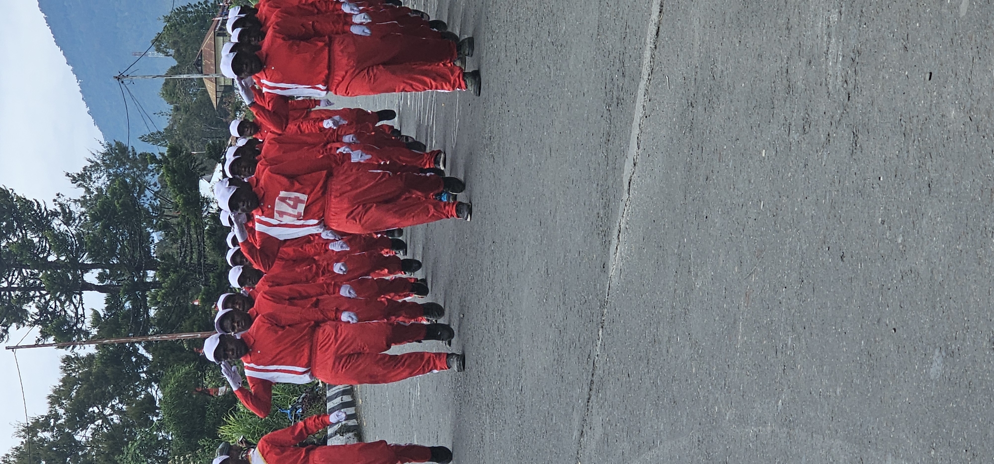 In the city of Puncak Jaya, Indonesia, a group of young people in red uniforms with white stripes are marching in formation on a paved road. They are all wearing white caps and some have white gloves. The person in the foreground is wearing a uniform with the number "14" in white on the chest. The background shows a street with trees and mountains in the distance, under an overcast sky.