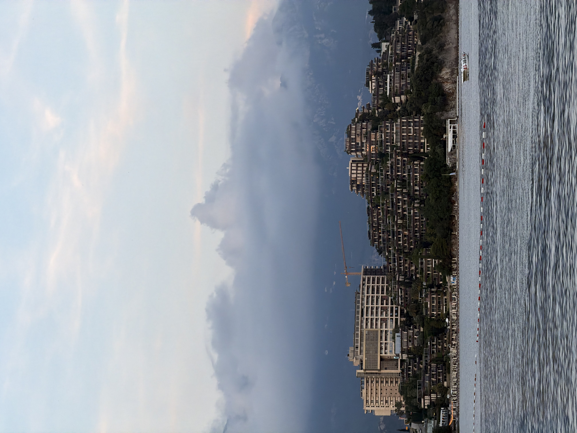 This image captures a vibrant coastal scene in Budva, Montenegro, likely during the late afternoon or early evening, characterized by dramatic weather conditions. The sky is divided, with the left side displaying a soft, pale blue with faint, wispy clouds, while the right side is dominated by a mass of dark, foreboding gray-blue clouds, suggesting an approaching or receding storm.
Below the dynamic sky, a densely developed coastline stretches across the frame. Numerous multi-story buildings, predominantly modern in design with many balconies, are terraced up a green hillside, indicating a popular resort or residential area. Interspersed among these structures are patches of vegetation and trees. A prominent yellow construction crane rises above one of the newer, larger buildings, signifying ongoing development in the area.
The foreground is dominated by a calm body of water, likely the Adriatic Sea, with visible ripples across its surface. Several small boats, mostly pleasure craft with white hulls, are docked or floating near the shore. A distinct line of cylindrical buoys, alternating in red and white, extends into the water, possibly marking a swimming zone or a boundary. A white structure, perhaps a small pier or building, is also visible at the water's edge. No individual people or visible text are clearly discernible.