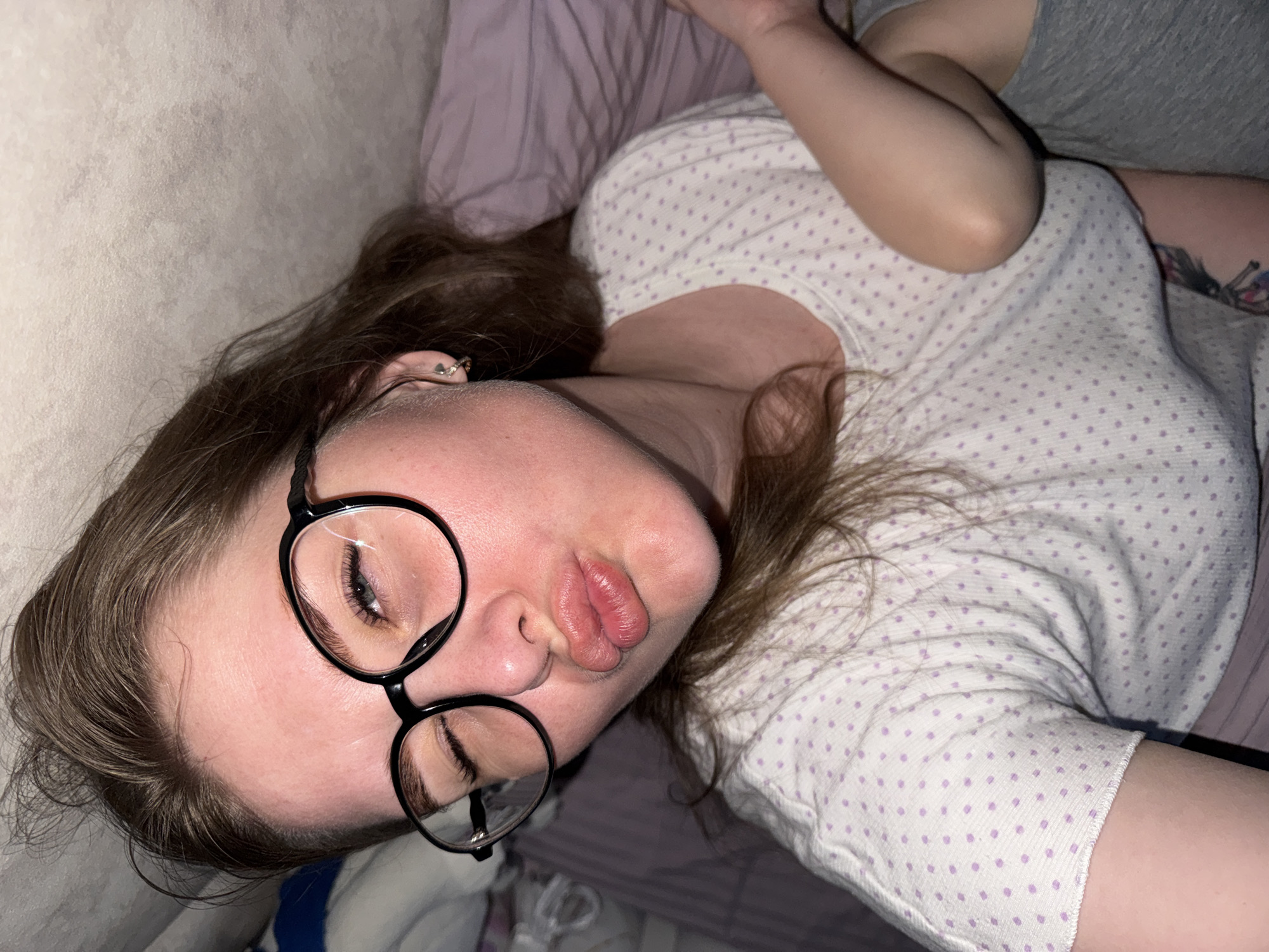 A young woman with long brown hair, wearing round black glasses, lies on a bed with a lavender pillow and a white shirt with purple polka dots. The woman is looking at the camera with her eyes closed, while pouting her lips. The location is implied to be in Gornyi (Dvurechenskoe S/P), Russia.