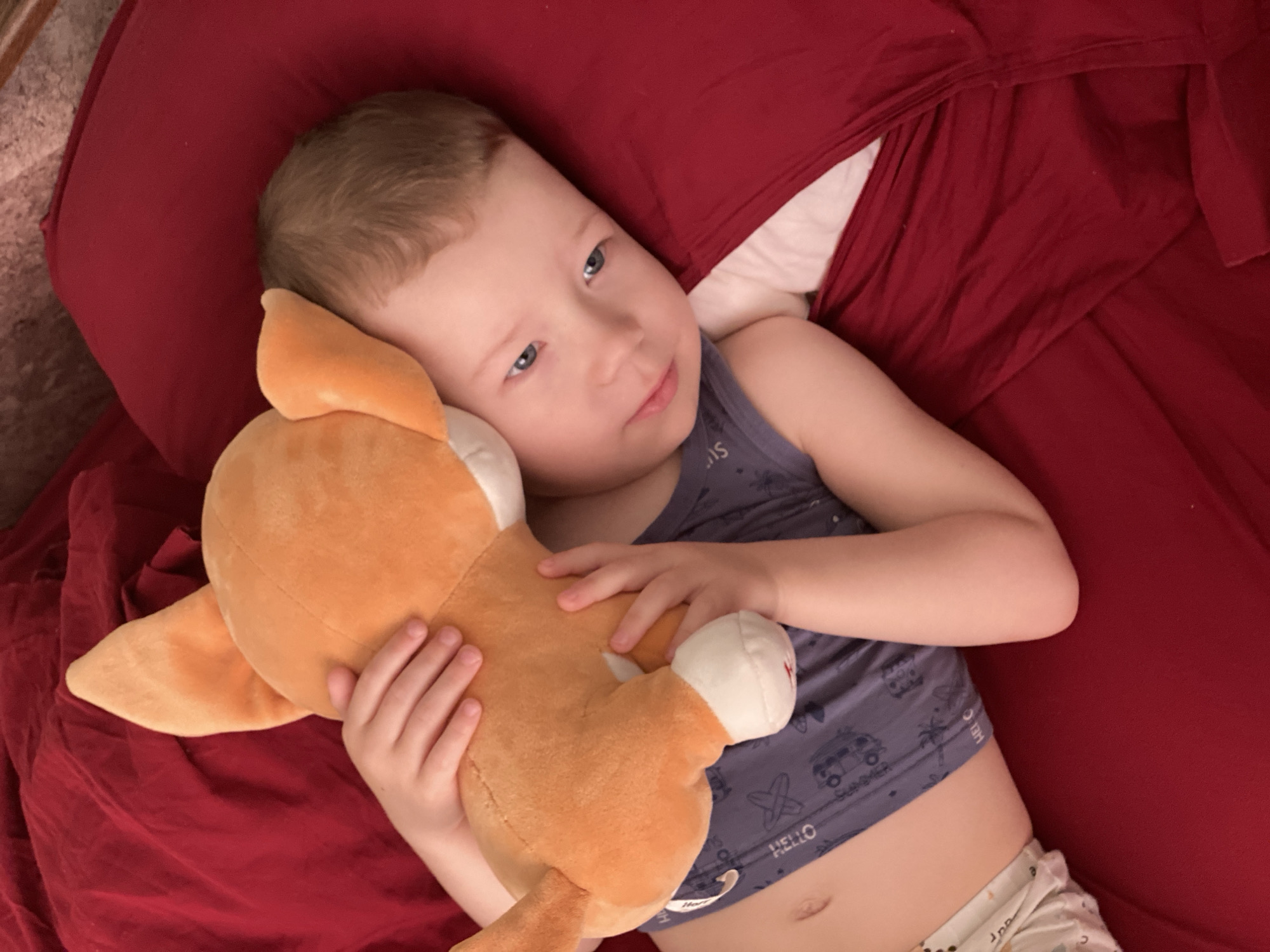 A young child, likely a boy of toddler or preschool age, lies on his back on a bed covered with rich red bedding. His head rests on a partially visible white pillow. With fair skin, light brown hair, and blue eyes, he gazes slightly upwards and to the right, wearing a soft, contemplative expression.
He wears a dark grey or blue sleeveless top patterned with light-colored symbols and text, including "HELLO," "Summer," palm trees, cars, and 'X' symbols. Against his chest, he gently cradles an orange plush toy, possibly a fox or dog, with white paws and a white belly patch. A small, indistinct red embroidered detail is visible on one of the toy's white paws. The child's lower abdomen, including his navel, and a portion of patterned undergarment or shorts are visible.
The scene suggests a quiet moment of rest or gentle play indoors. The red bedding, which fills most of the background, creates a warm, cozy atmosphere. The lighting appears soft and even, but the specific time of day is not discernible. No explicit outdoor weather or location cues are visible in the image.