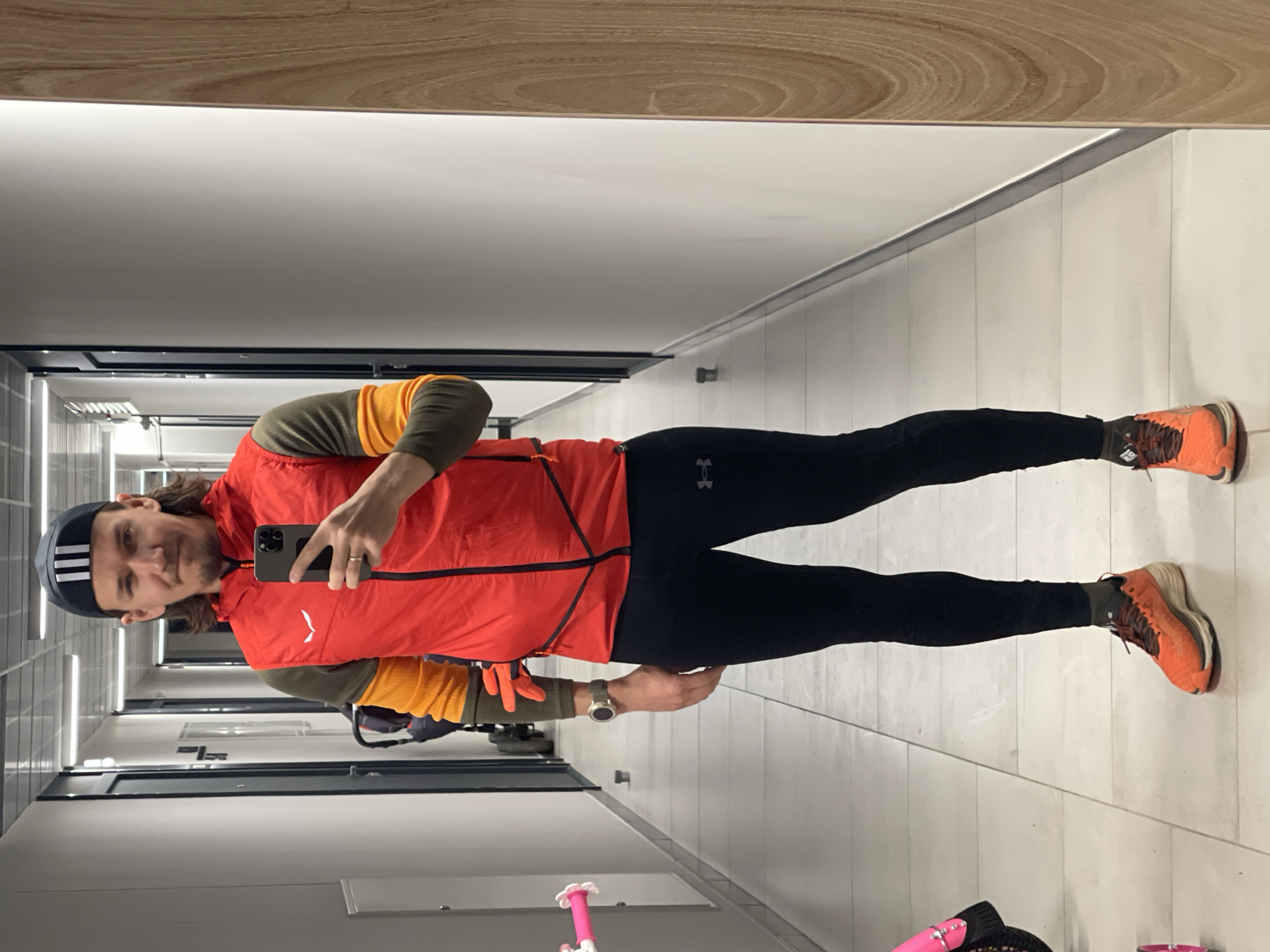 A man stands in a hallway, likely indoors, taking a selfie in a mirror. He wears a dark blue Adidas cap with white stripes, a red vest over a long-sleeved shirt with yellow and green portions, black leggings, and orange running shoes with brown laces. He holds a smartphone in his right hand, and a watch is visible on his left wrist. The hallway has light grey walls, tiled floors, and a light wooden trim near the ceiling. There are doors visible along the hallway. A pink child's scooter is also visible at the bottom right corner of the mirror.
I cannot specify the location as Izhevsk, Russia, as there is no evidence in the image to suggest this.