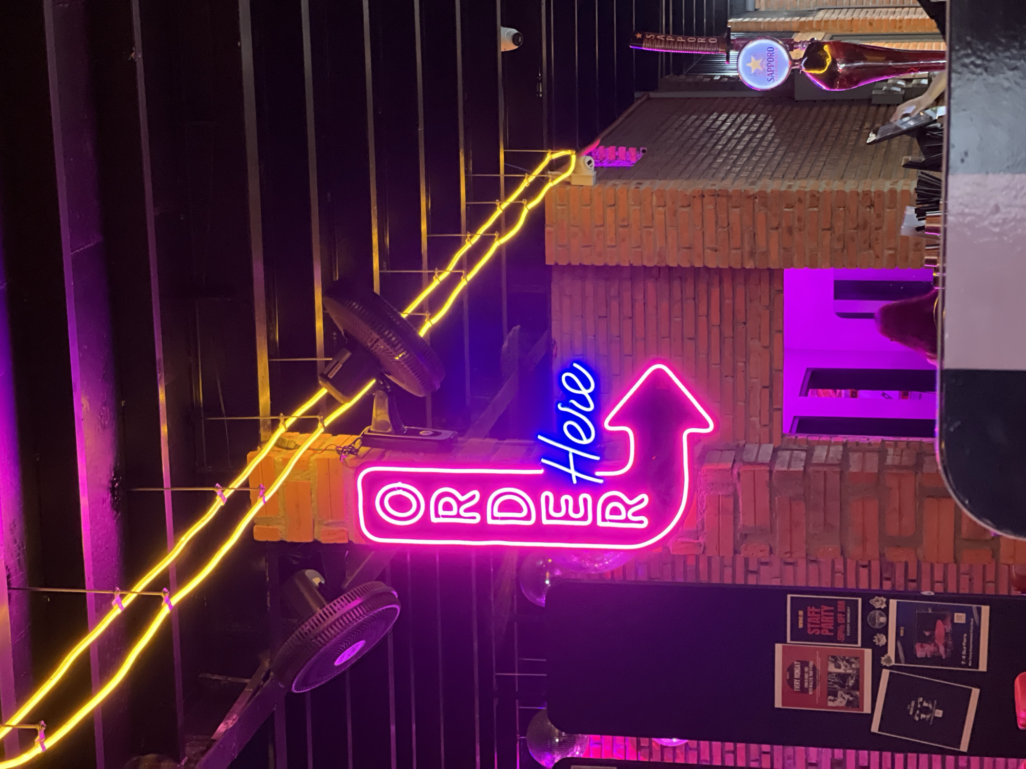 The image shows an illuminated "Order Here" neon sign in a business located in Bac Nha Trang Ward, Vietnam. The arrow sign is in pink and blue, positioned on a wall constructed of bricks and cinder blocks. A Sapporo beer tap is visible on top of this wall, along with posters and advertisements attached to the wall underneath the sign. The area is illuminated by various neon lights, with a fan positioned on the wall beside the sign. An intricate network of wiring can be seen going along the top left and continuing to the right of the frame. Metal support beams are also present in the background.
