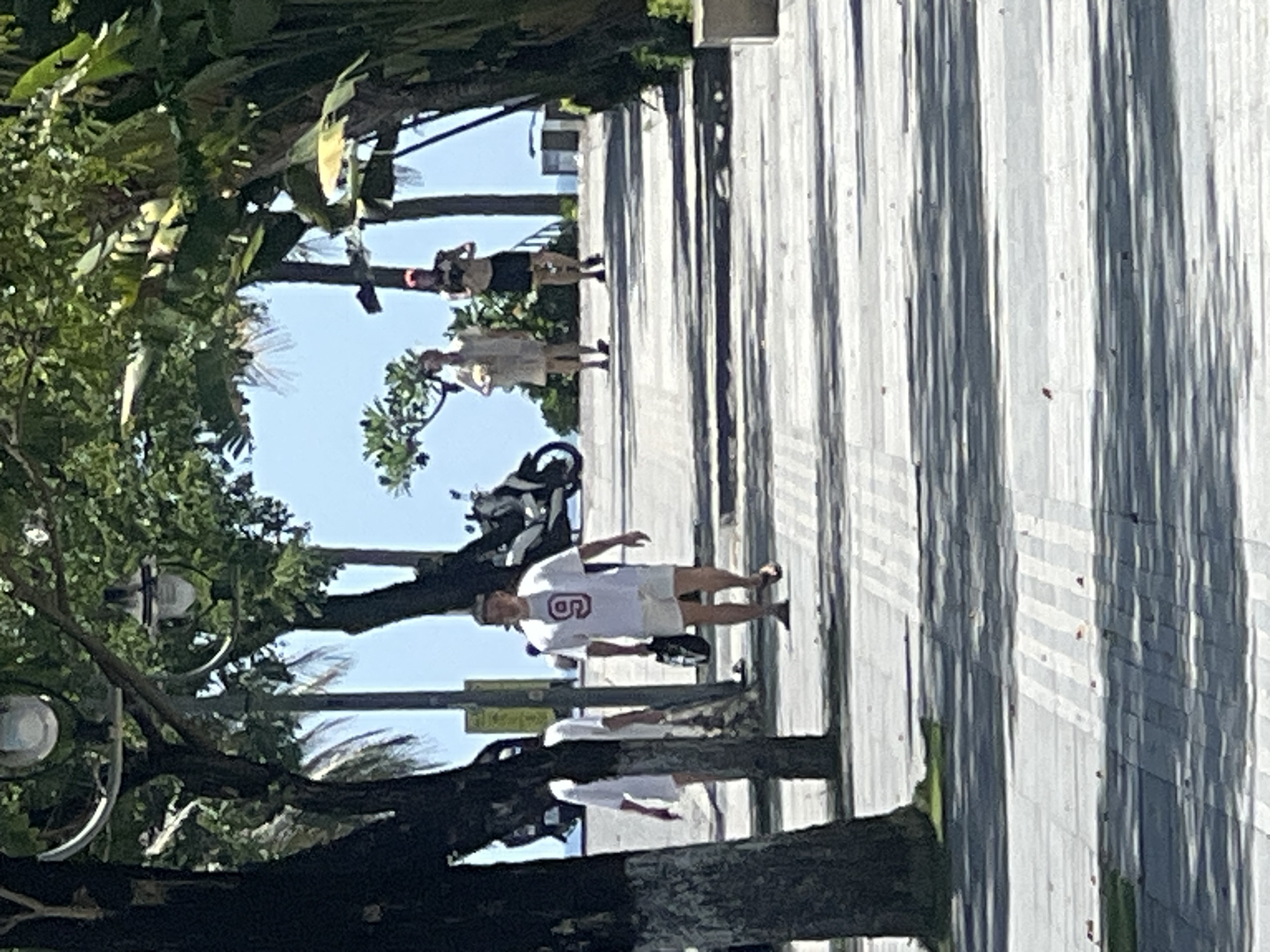 In Đà Nẵng, Vietnam, a paved walkway lined with trees and tropical foliage is depicted. The walkway is partially shaded by the trees, creating a pattern of light and shadow on the ground. Several individuals are visible walking along the path. In the foreground, a man wearing a white t-shirt with a red number "9" and white shorts is walking towards the camera, carrying a bag. Behind him, another person in a white t-shirt and shorts is walking away from the camera. Further up the path, two more individuals are seen walking. A scooter is parked to the left, and a glimpse of the ocean and sky is visible in the background. The overall scene suggests a sunny day in a pleasant outdoor environment.