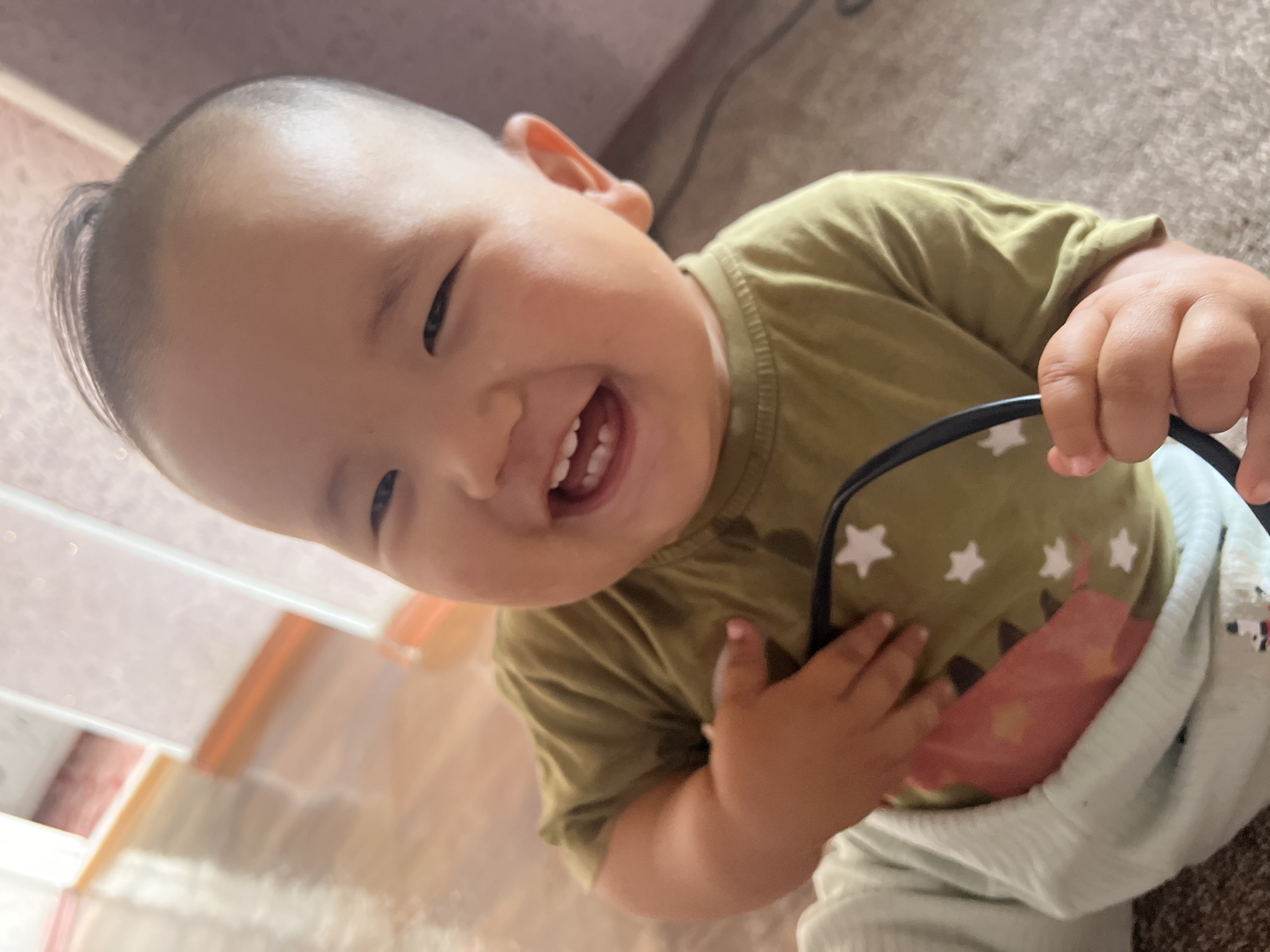 The image shows a close-up of a young child, likely a toddler, with a wide, joyful smile. The child has dark hair styled with a small tuft on top, and appears to be wearing a green t-shirt with a star pattern, and light-colored pants. The child is holding a dark, round object, possibly sunglasses or a toy, in their right hand. The background is blurred, showing a wooden floor and what appears to be an indoor setting with soft lighting. There are no clear indications of the specific location within Kara-Dıykan, Kyrgyzstan, as the image is focused on the child and does not contain any recognizable landmarks or environmental features that would pinpoint its exact location within the city.