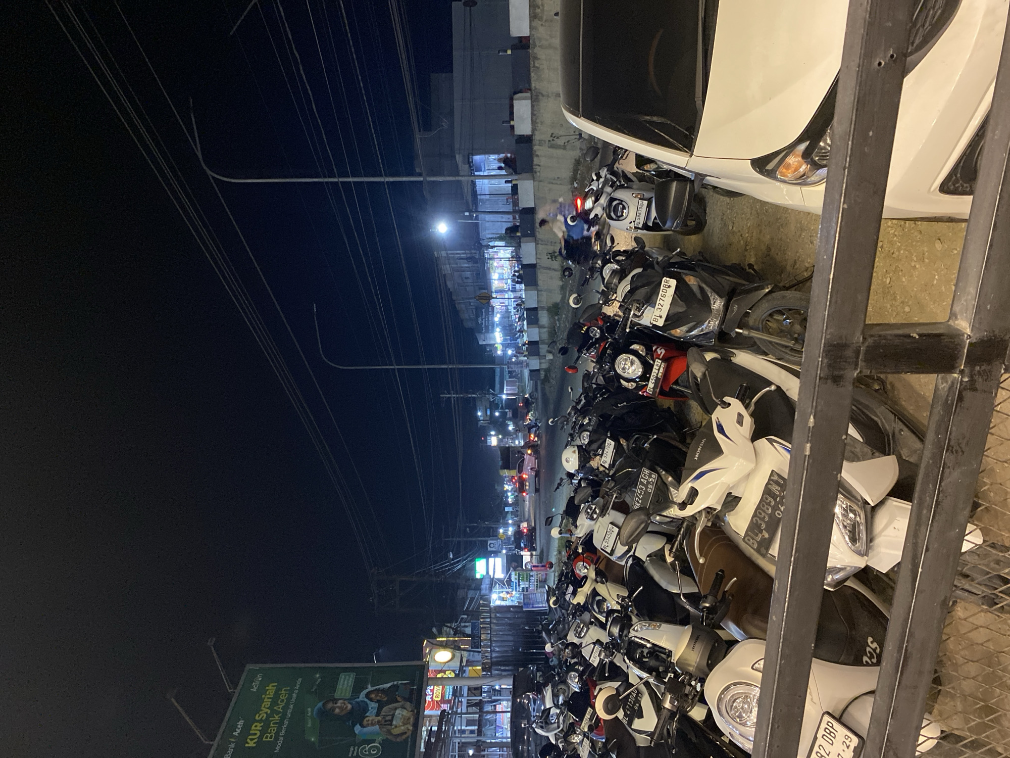 At night in Seuneubok Teungoh, Indonesia, a dense row of parked motorcycles, including many Honda scooters, dominates the foreground, stretching along the edge of a street near a white car and a metal railing. A billboard advertising "KUR Syariah Bank Aceh" is visible. Streetlights illuminate a row of shops and buildings in the distance. Power lines are strung overhead. The general setting is that of a busy urban street.