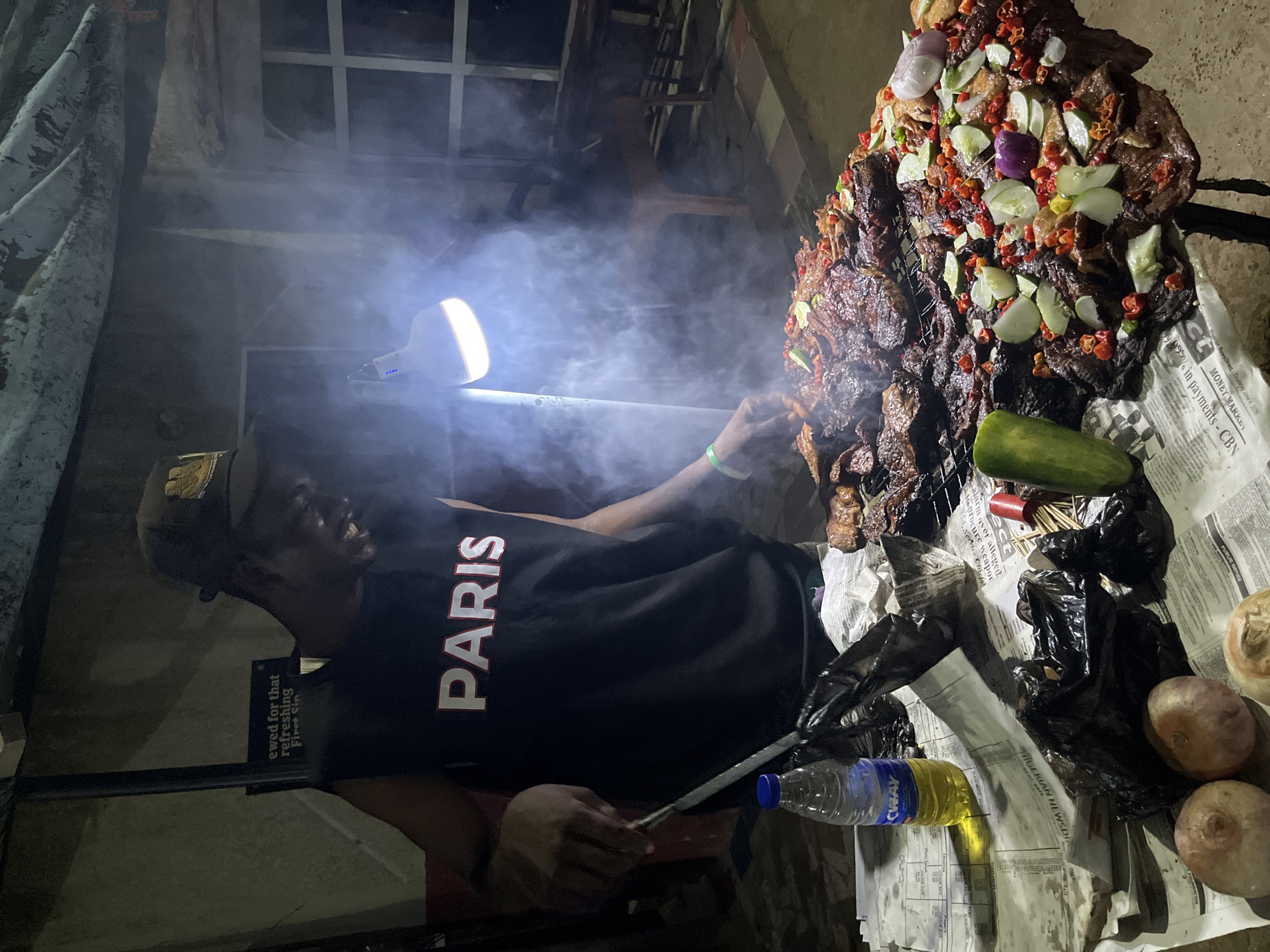 In Abuja, Nigeria, a street food vendor, a male, wearing a dark sleeveless shirt with "PARIS" printed on it in white, and a dark cap, is preparing grilled meat on a roadside stall. The scene is illuminated by a bright portable lamp, casting a glow and creating a smoky atmosphere. The vendor is actively tending to the meat on a metal grill using a large metal spatula. A water bottle with a blue cap and yellow liquid, along with several plastic bags, are on the table in front of him, partially covered by newspapers. The grilled meat, some of which appears to be in large pieces and others in smaller, more seasoned portions, is laid out on the grill and on the table, garnished with sliced cucumbers, red chili peppers, and red onion pieces. Nearby, raw onions are visible on the table. The background shows a dimly lit outdoor environment with corrugated roofing and what appears to be part of a structure. The overall impression is a bustling night market scene.