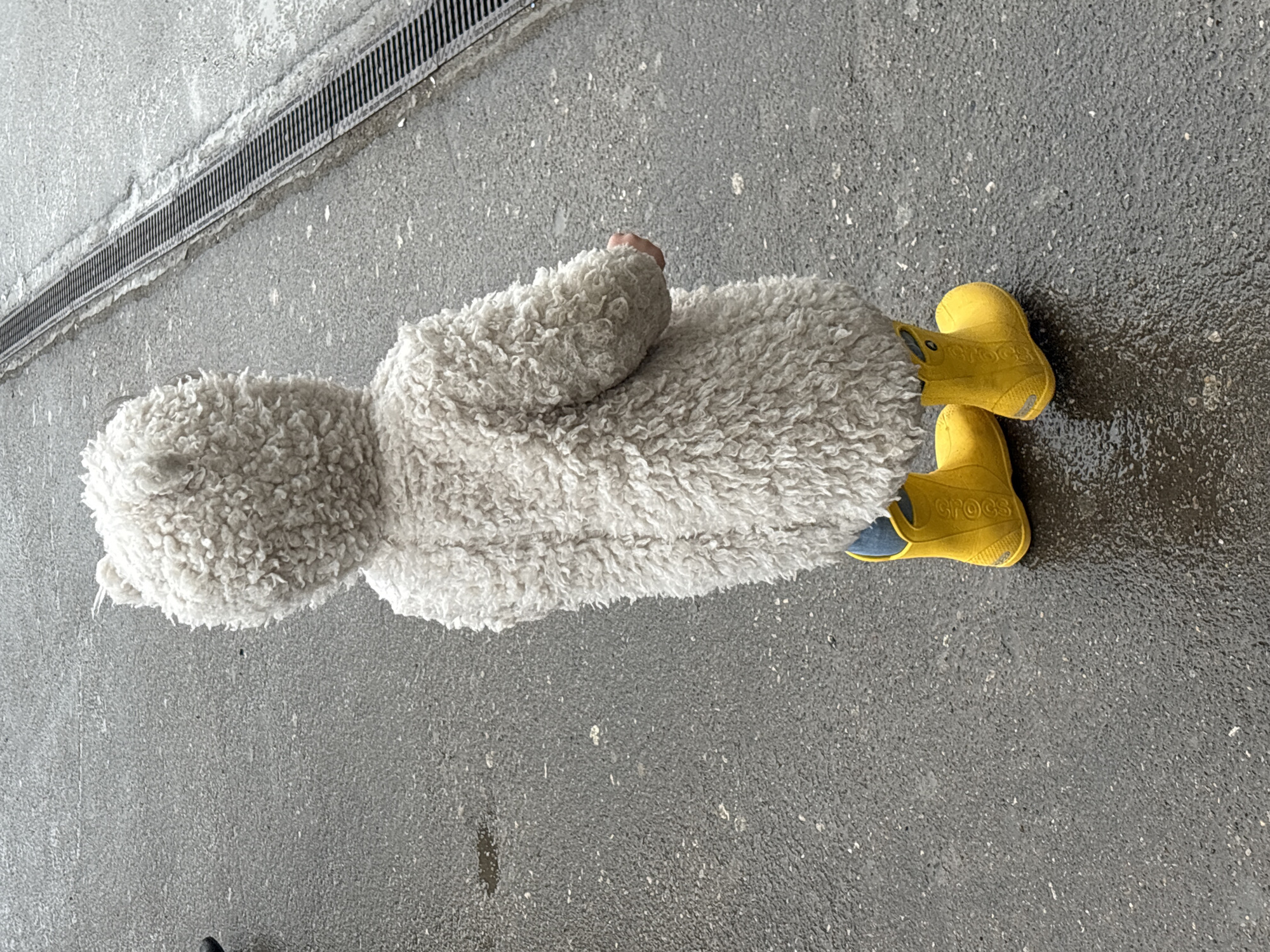 A child wearing a white, fleece-like jacket and yellow Crocs stands on grey asphalt, seemingly outdoors. A metal grate is partially visible near the child. No further information is available to determine the location within Perm, Russia.