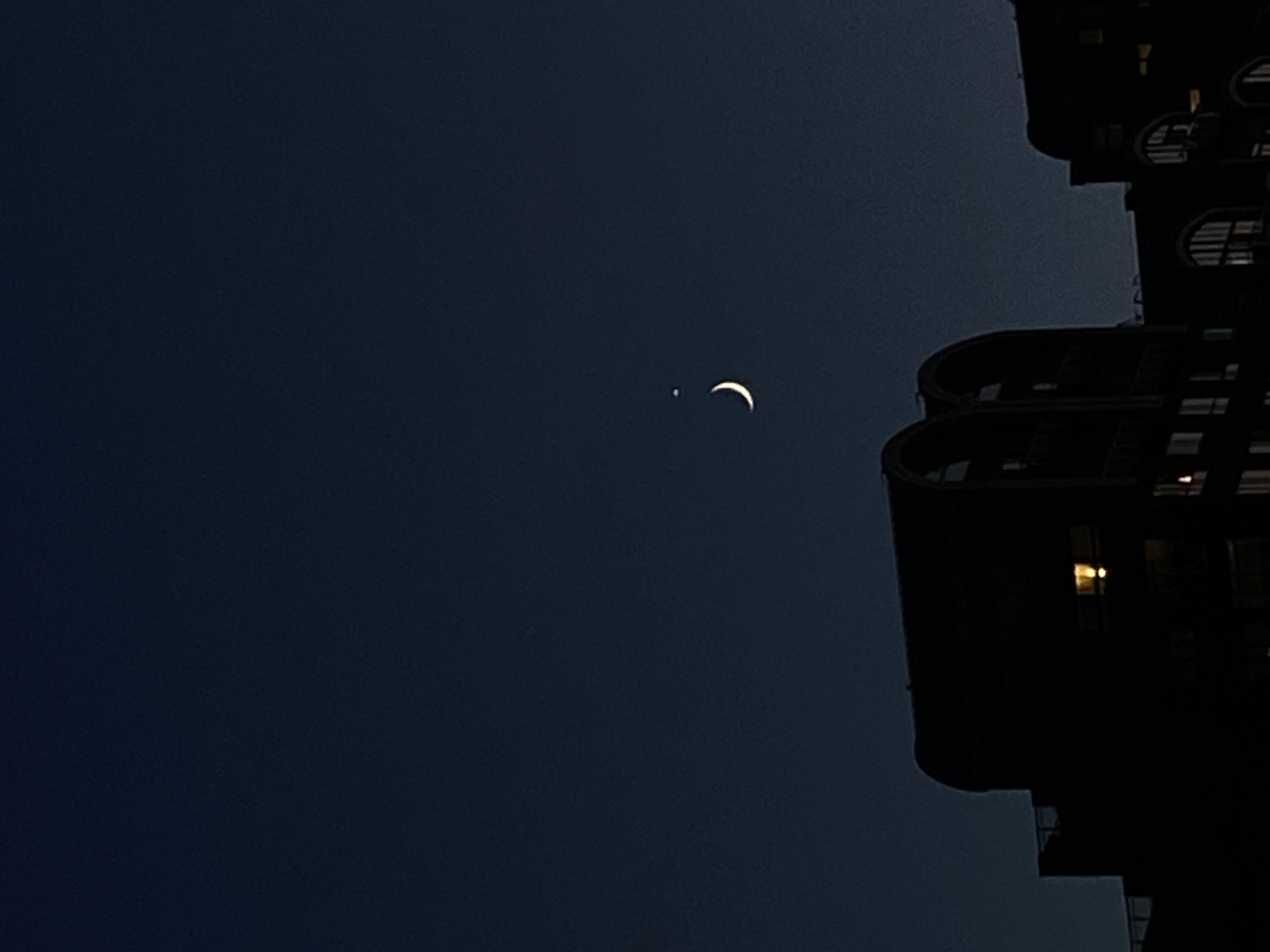 The image captures a nighttime scene in London, United Kingdom. Silhouetted against a dark blue, clear sky are the tops of several buildings with arched windows and balconies. In the upper center of the sky, a crescent moon is visible, accompanied by a small, bright star. One of the buildings in the foreground has a lit window, casting a warm glow into the darkness. The overall impression is one of urban quietude under a celestial display.