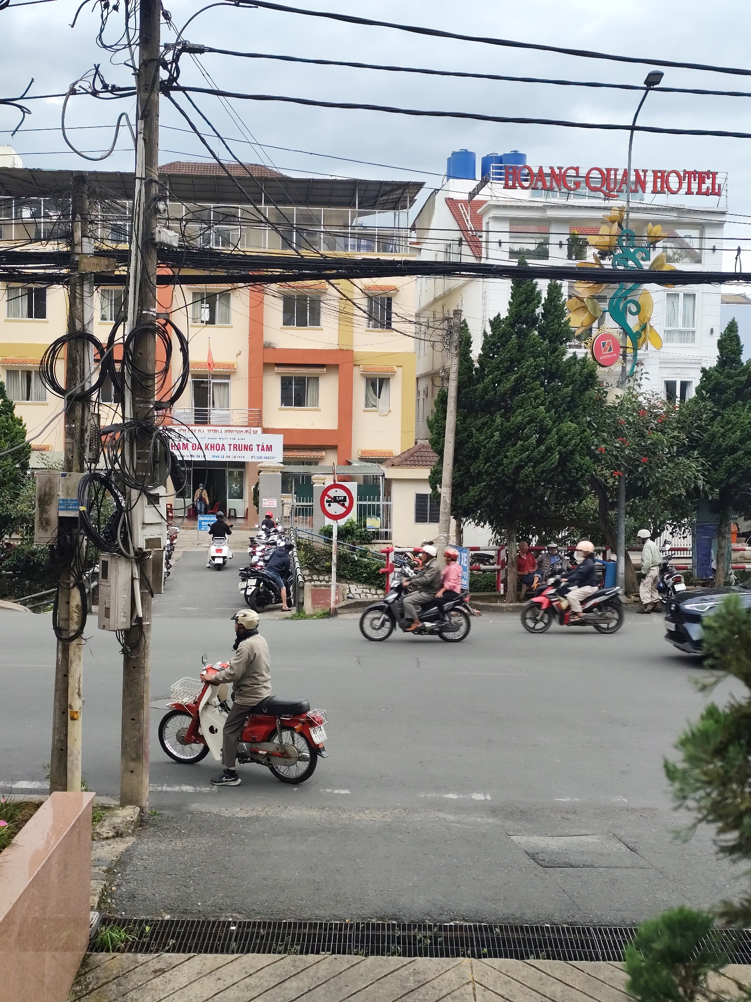 The image captures a street scene in a city. In the foreground, a man on a red and white motorcycle is facing away from the camera, moving to the left. The motorcycle has a white basket on the front and a license plate. The man is wearing a light-colored jacket and gray pants, with a light-colored cap on his head. To the left of the motorcycle, there are two utility poles with a dense network of black electrical wires. A concrete wall with a pinkish-beige finish is visible in the lower left corner, leading to a metal grate drain.
In the background, the street is busy with traffic. Several other motorcycles are moving in the same direction, some with riders wearing helmets and casual clothing. A white scooter is also present. Across the street, buildings line the road. A yellow and orange building with multiple windows and balconies is prominent, with a sign that reads "PHẨM DA KHOA TRUNG TÂM". Further down the street, a white building with the red sign "HOÀNG QUÂN HOTEL" is visible, topped with blue water tanks. Green trees are interspersed between the buildings. A traffic sign with a red circle and a horizontal bar indicates no entry. The sky is overcast and cloudy. The overall atmosphere appears to be a typical day in a bustling urban environment.