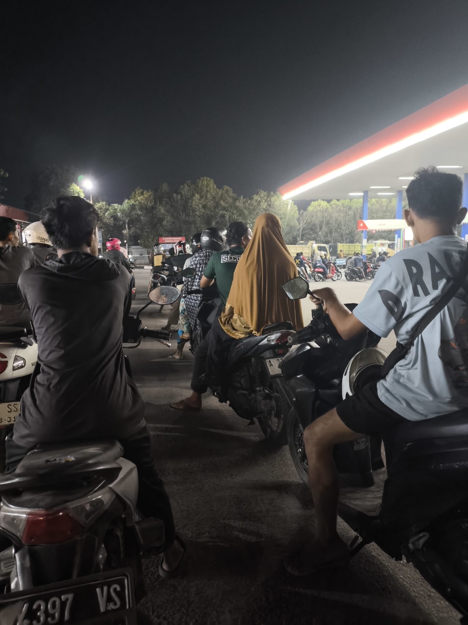 The image shows a nighttime scene at a gas station in Waringinkurung, Indonesia. A queue of motorcycles and their riders are waiting. The gas station canopy is illuminated with white and red lights. In the foreground, two individuals are seated on motorcycles. The person on the left is wearing a dark grey long-sleeved shirt, and the person on the right is wearing a light blue t-shirt with black lettering and black shorts. Further back, another individual is covered in a tan-colored headscarf and robe, seated on a motorcycle. Other people and motorcycles are visible in the background, along with trees and the dark sky. The ground appears to be paved.