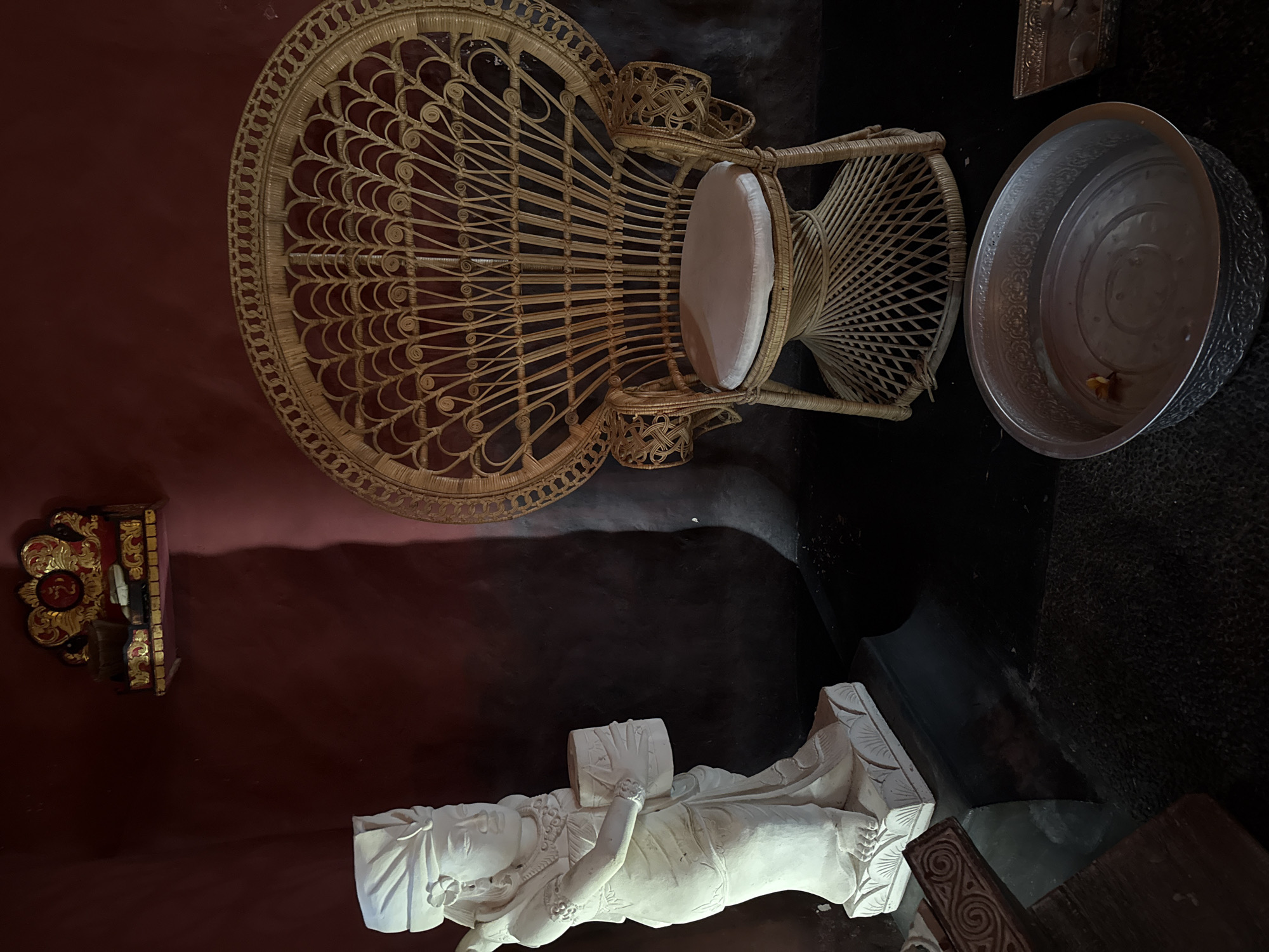 The image depicts a dimly lit, indoor scene that appears to be a shrine or a religious space, likely located in the Ubud District of Indonesia, given the artistic style and cultural motifs. Dominating the right side of the frame is a large, ornate peacock-shaped chair crafted from woven rattan. The chair has a high, rounded back featuring intricate patterns resembling peacock feathers and a cushioned seat. To the left of the chair stands a life-sized, white statue of a woman, possibly a deity or a significant cultural figure, adorned with traditional attire and jewelry, holding a cylindrical object in her hands. The statue is placed on a pedestal with decorative carvings.
In the foreground, a metallic, ornate bowl filled with water and a single flower petal rests on a dark surface, suggesting a ritualistic purpose. Behind the statue, on the maroon-colored wall, a small, gilded altar or shrine is mounted, adorned with red and gold embellishments and what appears to be a religious symbol. The overall ambiance is one of reverence and cultural significance, with shadows playing a crucial role in highlighting the forms and textures of the objects. The floor is dark and textured, possibly made of stone or concrete. The lighting is atmospheric, creating a sense of depth and mystery.