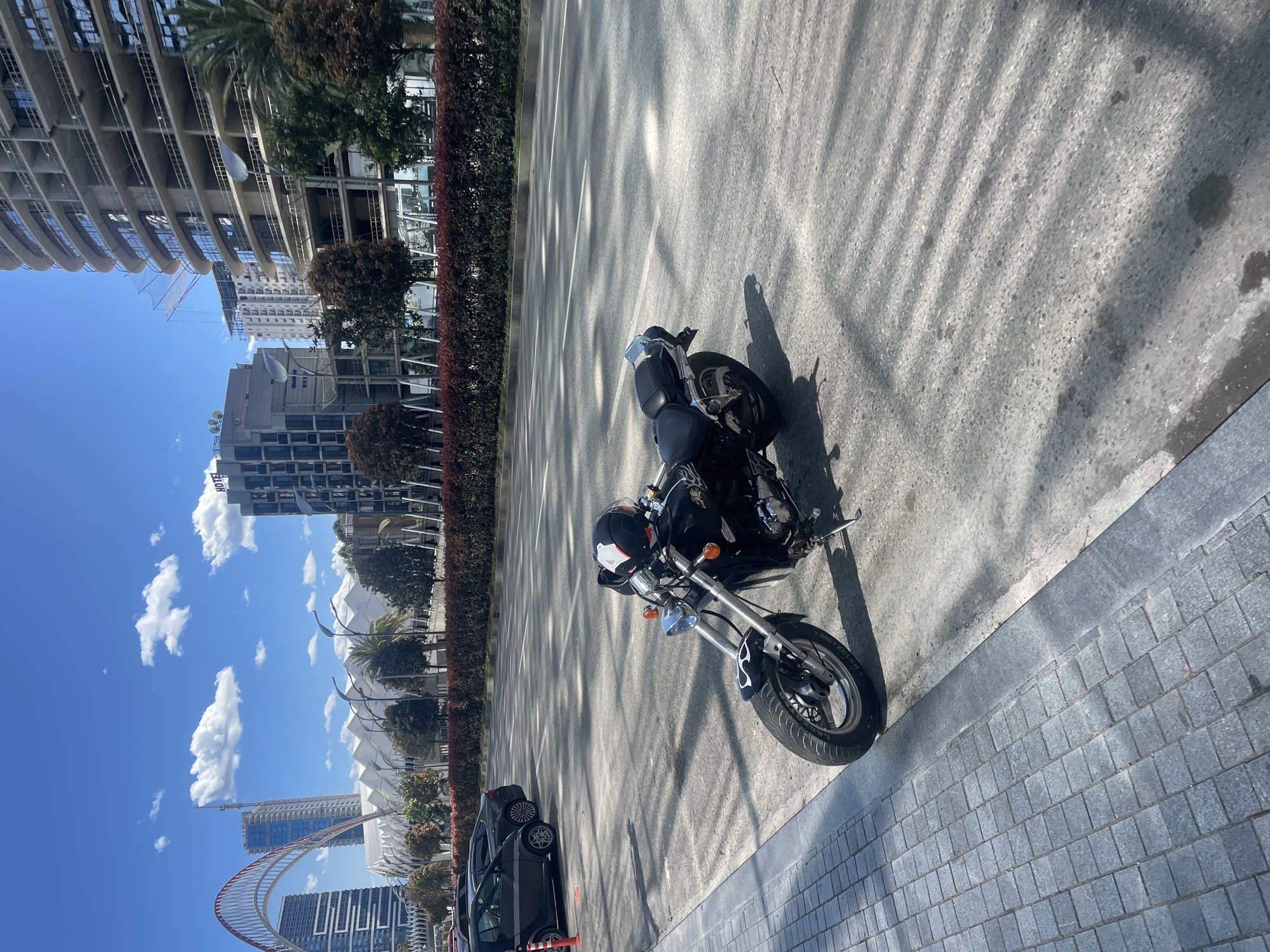 The image shows a motorcycle parked on a paved road next to a sidewalk in Batumi, Georgia. The sky is blue with scattered clouds. A row of bushes lines the road. Buildings and a red arch structure are visible in the background. A black car is parked nearby. The motorcycle is black with a chrome front fork and a white-and-black helmet rests on the seat. The ground is light grey and shows shadows of nearby structures.