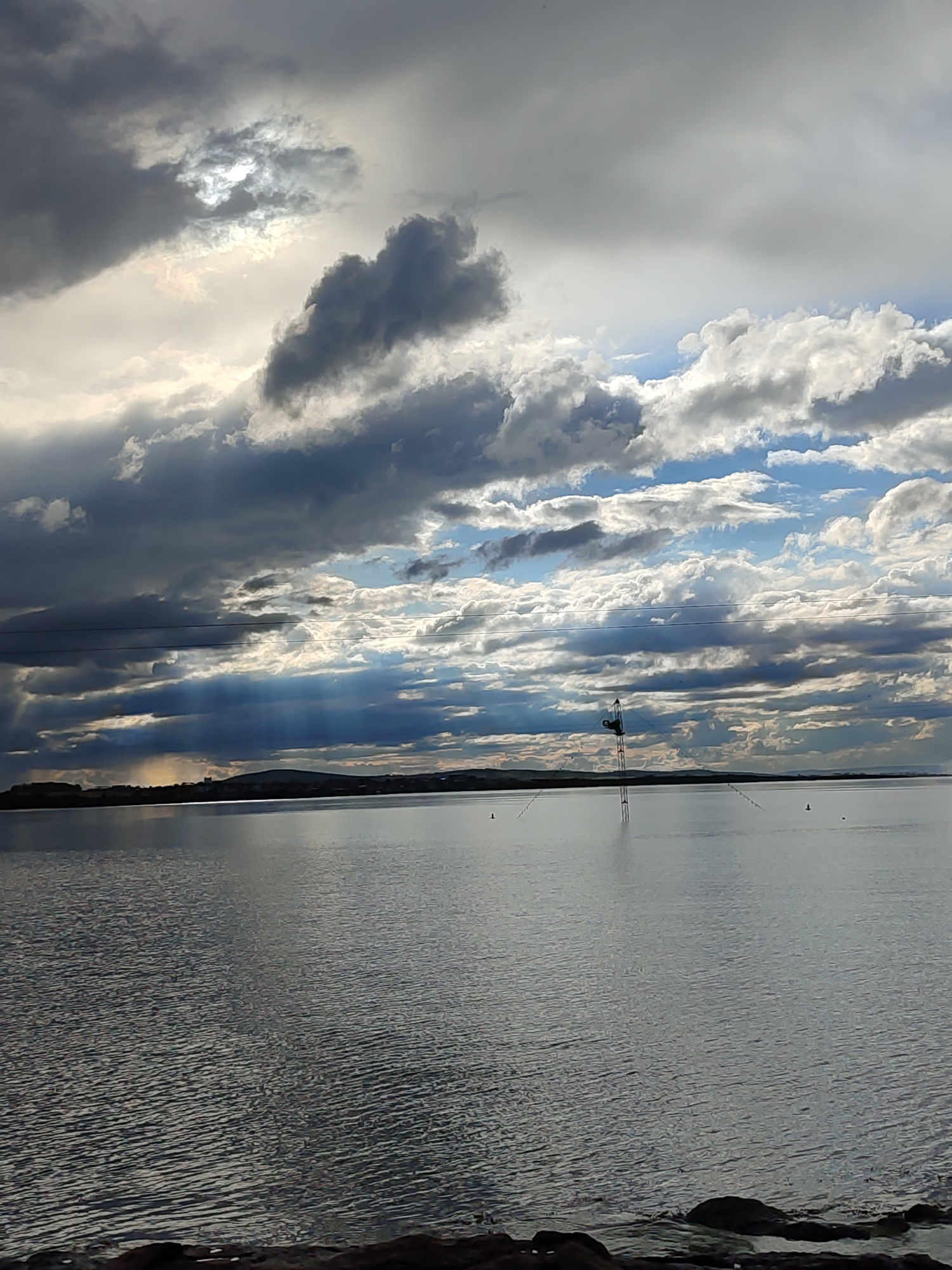 The image depicts a body of water, likely the Black Sea, in Burgas, Bulgaria, under a dramatic sky. The sky is filled with a mix of dark, storm-like clouds and patches of brighter, sunlit clouds. The sun is partially obscured by the clouds, creating a diffused, ethereal glow. The water reflects the overcast sky, appearing grey and rippled with slight wave action. In the distance, there is a coastline with a low, undulating land mass. In the middle of the water, there is a tower-like structure, possibly for wakeboarding or water sports, with cables extending to buoys in the water. Some wires run above and behind. The foreground shows the shoreline with dark rocks or stones.
