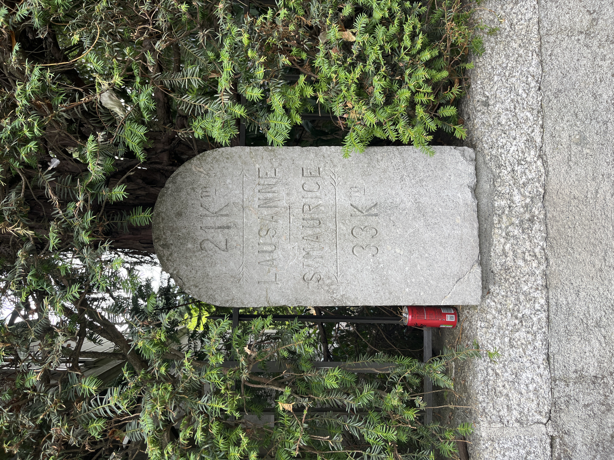 A rough-hewn, vertical stone marker, weathered and inscribed, stands partially obscured by lush green conifer branches. The marker is positioned in front of a dark metal fence and is situated on a granular stone wall. The top of the marker is rounded, and it is inscribed with three lines of text. The first line reads "21 K" with decorative lines above and below the "K". The second line reads "LAUSANNE". The third line reads "S. MAURICE" with a decorative arrow pointing to the right. The fourth line reads "33 K". To the left of the marker, a red soda can is visible, resting against the stone wall near the base of the fence. The background shows further foliage and the suggestion of a building. The scene is located on the Quai de Montreux in Montreux, Switzerland, adjacent to the Chillon Castle.