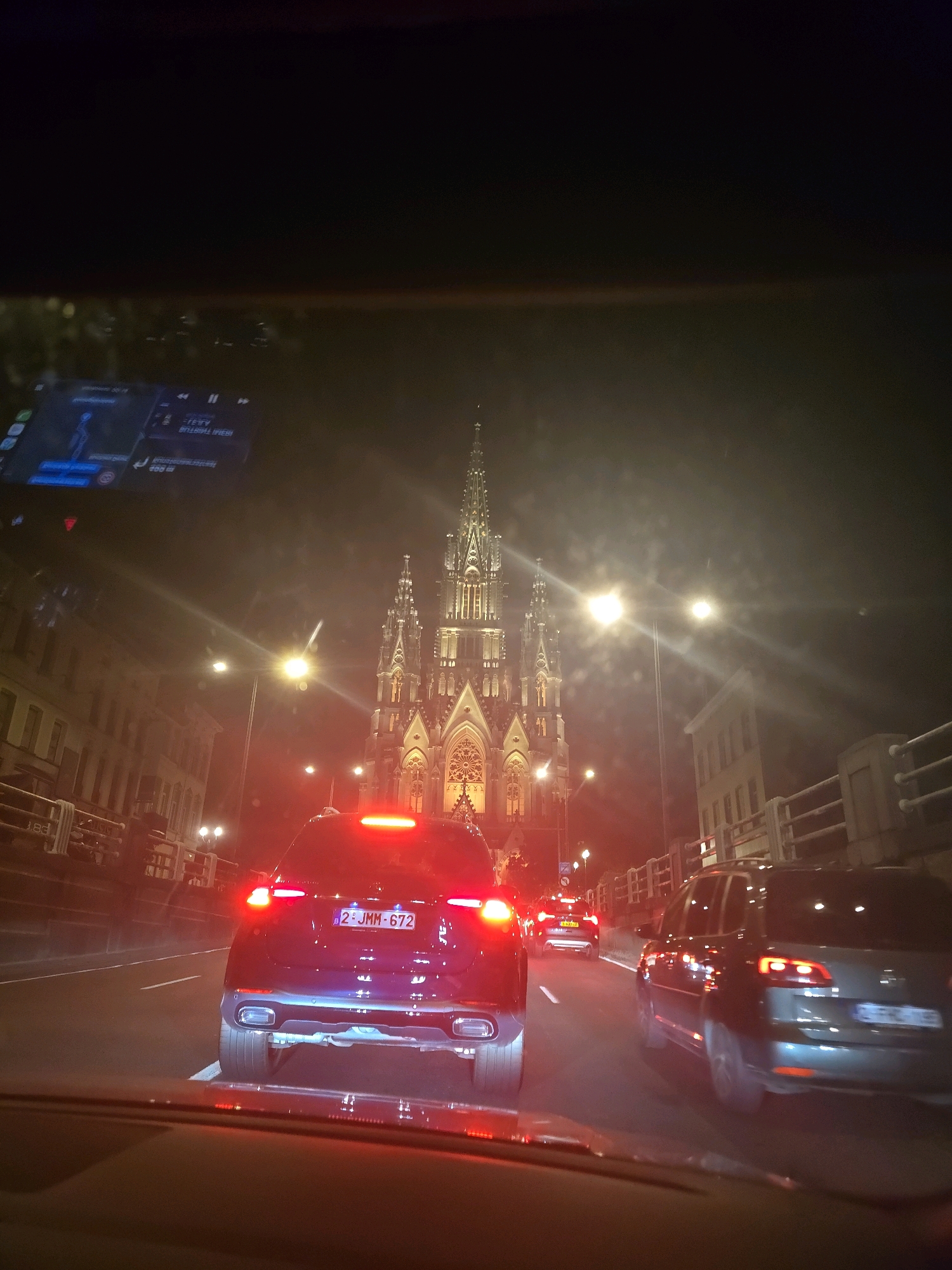 A nighttime view from inside a vehicle shows traffic on a street in front of a large, illuminated Gothic-style church. The church, with its tall spires and ornate facade, is the focal point of the background. Streetlights and building lights cast a warm glow on the scene. In the foreground, the taillights of several cars are visible, indicating a queue of vehicles. The car in the immediate foreground has its brake lights illuminated, and its license plate reads "2 JMM 672". The overall atmosphere is urban and serene, with the majestic church serving as a prominent landmark. This scene is likely in Brussels, Belgium, given the architectural style of the church and the presence of a European license plate.