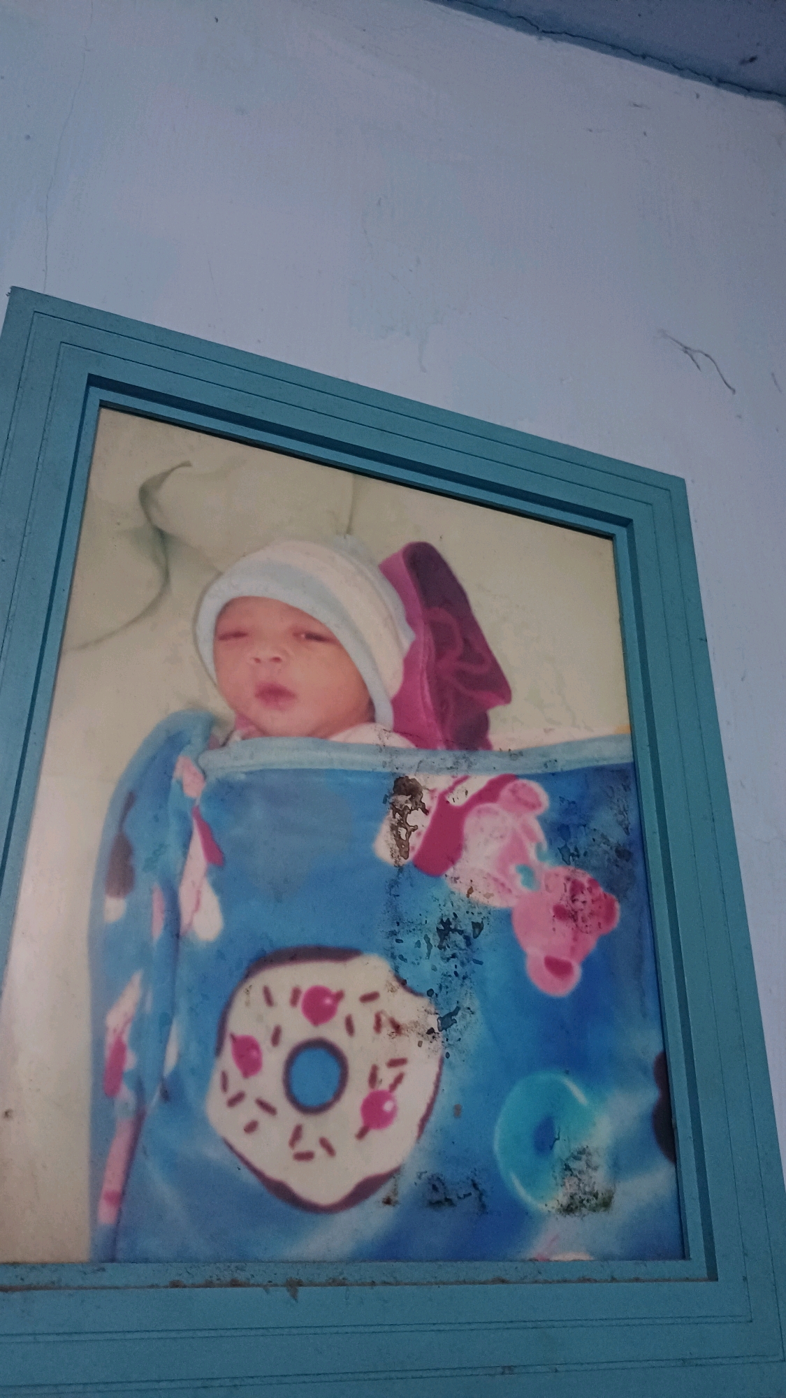 A framed photograph of a newborn baby wrapped in a blue blanket with donut and cartoon character patterns is hanging on a light blue wall. The baby is wearing a white and blue striped hat and appears to be asleep or drowsy. The photo is dusty and slightly damaged. The wall has some visible cracks and texture. The location is specified as Kowangan, Indonesia.
