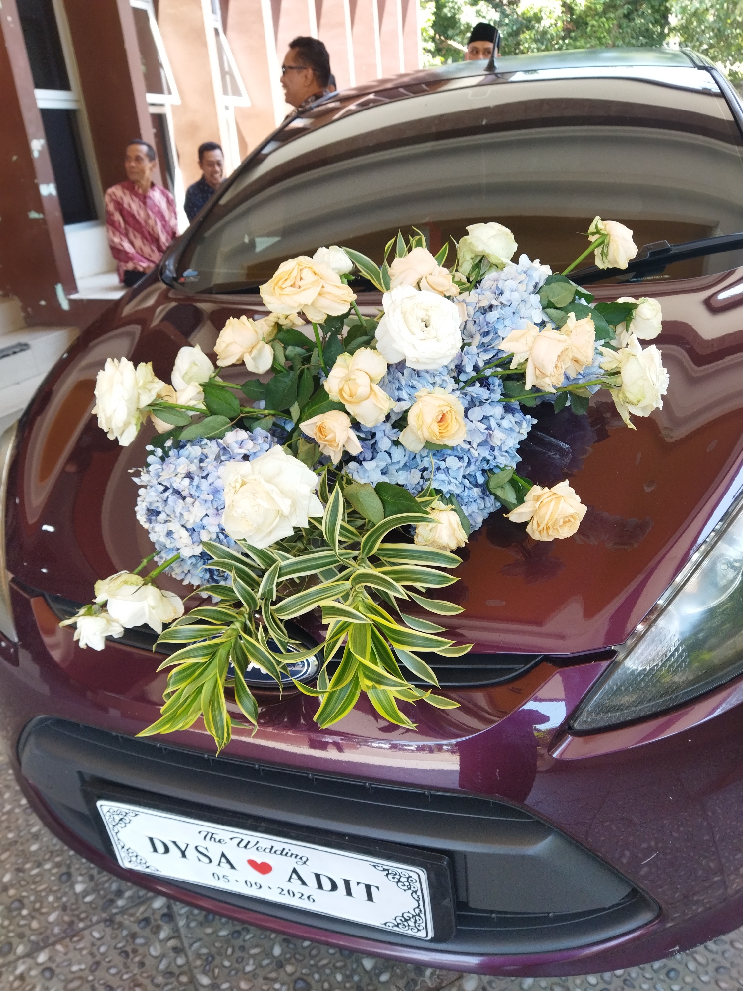 A maroon sedan is parked on a tiled surface in front of a building. The hood of the car is adorned with a floral arrangement featuring light yellow and white roses and light blue hydrangeas, along with green and yellow striped leaves. A white license plate with black lettering is affixed to the front of the car, displaying "The Wedding DYSA ❤️ ADIT 05 - 09 - 2026." In the background, three men are visible. One man, wearing glasses and a patterned maroon and white shirt, stands to the left, looking away from the camera. Another man, wearing a dark shirt with a pattern, is in the middle background, also looking away. A third man, wearing a black cap, peeks over the top of the car. The building in the background has a brown facade with large windows and cream-colored vertical accents. The scene appears to be outdoors with greenery visible in the upper right corner. The overall context suggests a wedding car decoration. The location is the city of Semarang, Indonesia.