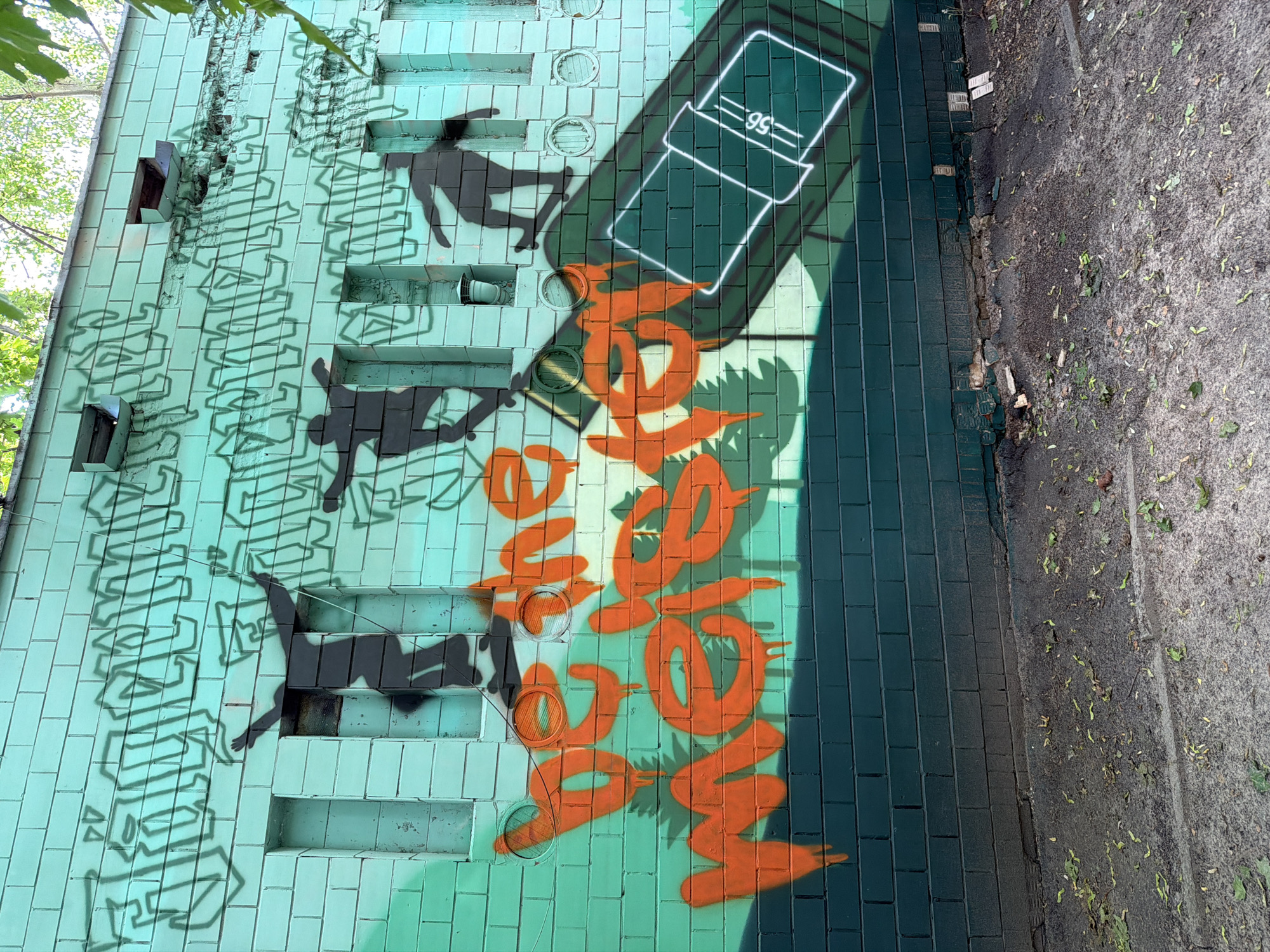 The image depicts a section of a building's exterior covered in graffiti, located in Kyiv, Ukraine. The wall is tiled with a light mint green color. On the left side, there are several shadowy silhouettes of people, some appearing to be falling or in dynamic poses. Faint, dark green graffiti lettering in a blocky, stylized font is visible across this section of the wall, partially obscured by the silhouettes.
To the right, there is a large, stylized drawing of a white rectangular object with a handle, resembling a simplified carton or box, outlined in bright white and containing the number "95" within a smaller rectangle. Beneath this object, large, bold orange graffiti letters spell out "LOVE" in a flowing, decorative script with sharp, pointed edges.
The building appears weathered, with some damaged tiles and exposed brickwork on the left. Several small, circular vents or openings are visible on the wall. The ground next to the building is a mix of dirt and asphalt, with scattered leaves, suggesting an outdoor urban setting. The lighting suggests it is daytime, with shadows cast by the trees on the upper left and by the graffiti elements. The overall scene is one of urban decay and artistic expression.