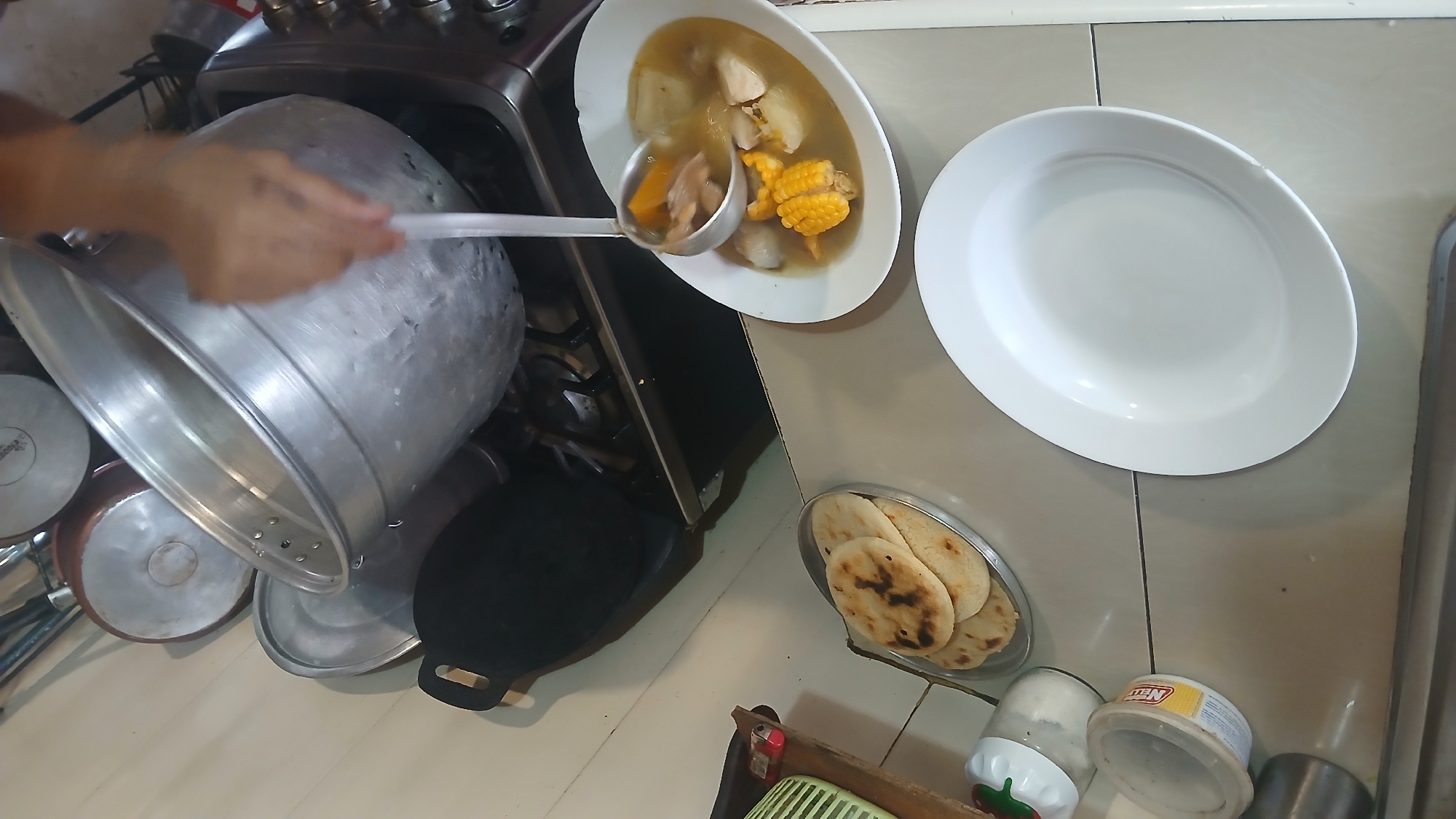 A person is using a ladle to serve sancocho, a traditional soup, from a large aluminum pot into an oval serving dish. The soup contains pieces of corn on the cob, squash, potatoes, and meat in a broth. Next to the serving dish is a large, empty white oval plate. Below the plate is a small, round silver dish with four arepas, a type of cornbread. Also visible are a stove, other cooking pots, and two plastic containers. The scene is set in a kitchen. While Altagracia, Venezuela, is mentioned, there are no elements in the photograph to confirm this location.