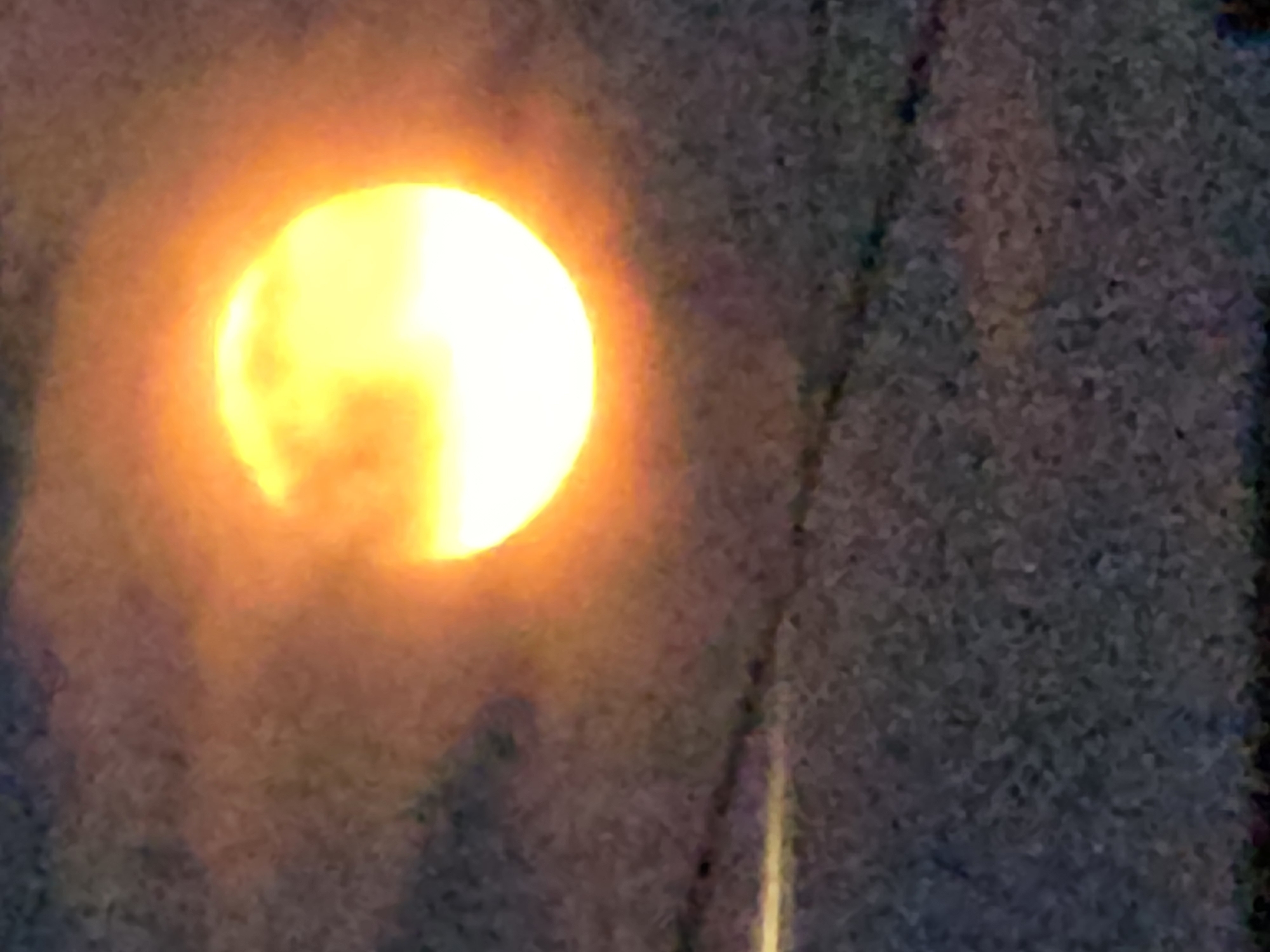 The image shows a partially obscured sun with a bright yellow and orange glow, set against a dark, textured background. The sun is located in the upper right quadrant of the image. Faint horizontal lines are visible in the lower portion of the image, suggesting the presence of power lines or similar structures. The overall scene appears to be outdoors, possibly during twilight or with atmospheric conditions affecting visibility. There are no people or identifiable objects other than the sun and implied foreground structures. The location is given as Буренка, Russia.