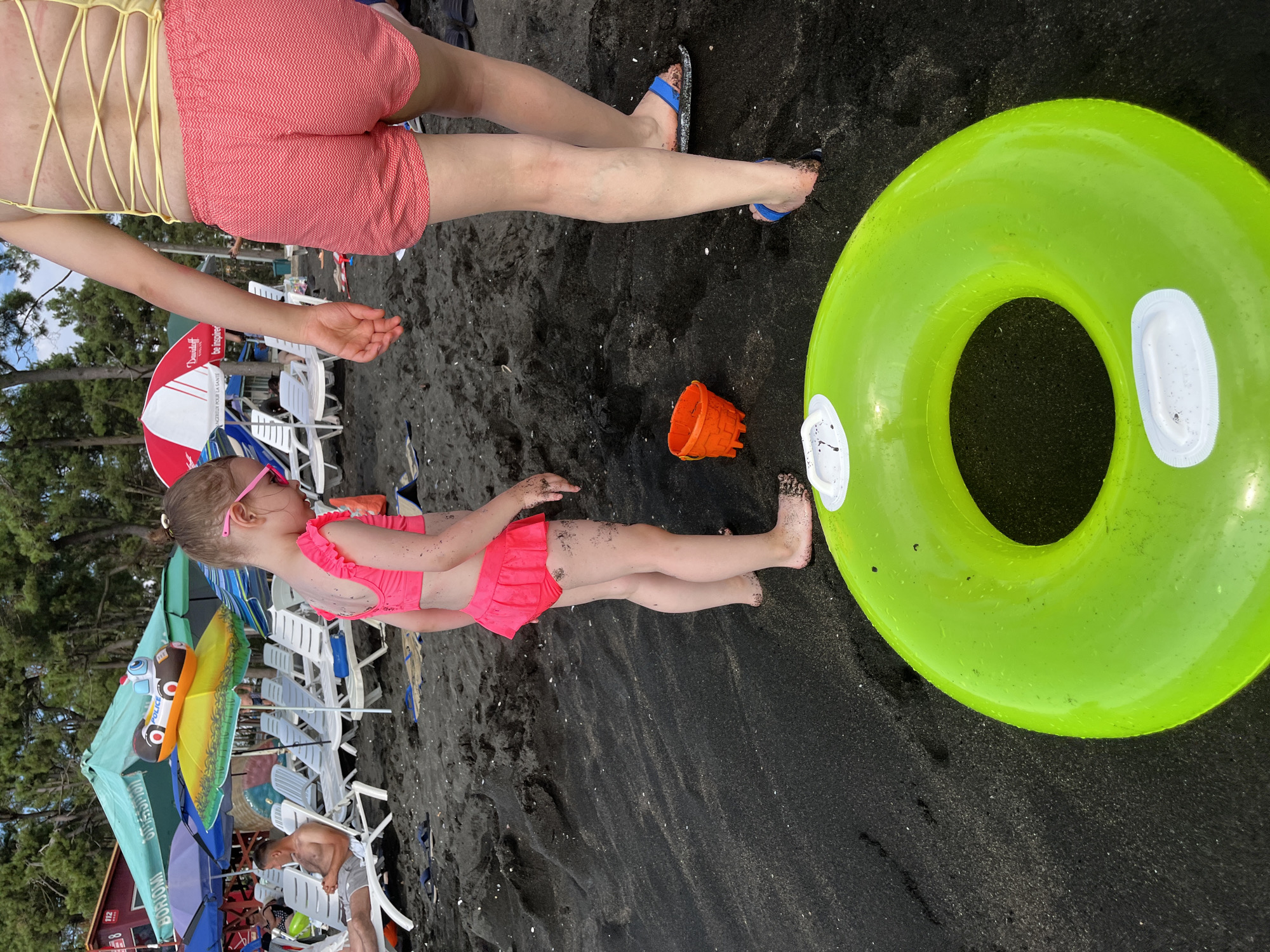 The image depicts a beach scene in Ureki, Georgia. The primary subjects are two individuals, an adult and a child, on black sand. The adult, with her back to the camera, wears a yellow bikini top with a crisscross back and coral-pink patterned shorts. Her legs are visible, with blue and black flip-flops on her feet. To her right, a young child in a pink ruffled swimsuit is seated and looking to the left, wearing pink sunglasses. In front of the child is a bright green inflatable ring. An orange plastic bucket lies on the sand between the child and the ring.
In the background, numerous white beach chairs are arranged under colorful umbrellas, suggesting a developed beach area. Trees are visible behind the chairs, indicating a coastal setting. The sand appears dark, consistent with the black sand beaches of Ureki. The lighting suggests it is daytime, and the presence of beachgoers and umbrellas implies fair weather. The overall mood seems relaxed and indicative of a day spent at the beach. Some text is partially visible on one of the umbrellas, but it is not legible enough to discern.