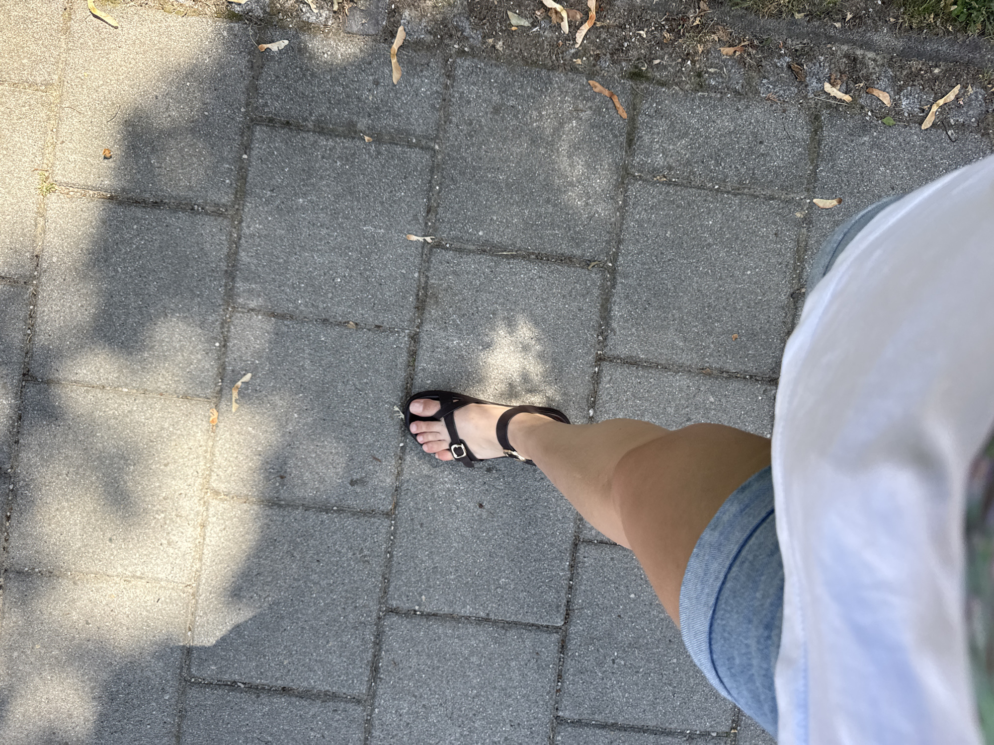 The image shows a person's right leg and foot, wearing a denim skirt and a black sandal, stepping on a paved walkway composed of rectangular gray concrete blocks. The walkway is partially shaded by tree branches, casting dappled shadows. Dried leaves and seeds are scattered on the walkway and along the grassy verge to the right. The scene appears to be outdoors on a sunny day. The location is specified as Höhenkirchen-Siegertsbrunn, Germany.