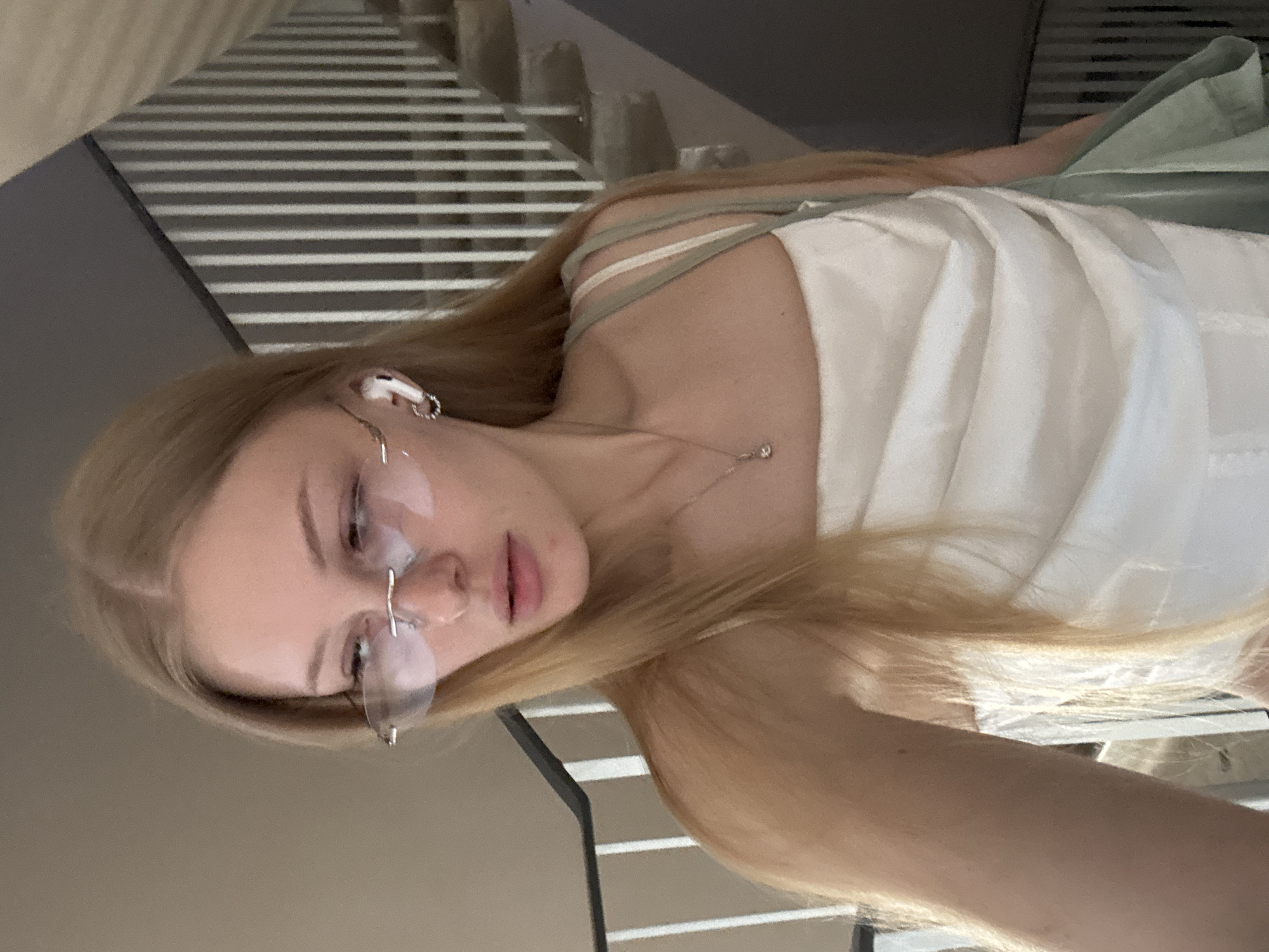 A young woman with blonde hair, wearing rimless glasses, a white strapless top, and wireless earbuds, is standing on a staircase. She is looking down and to her left. A light green bag is draped over her left shoulder. The staircase is made of concrete with metal railings. The background consists of a wall and a railing. The lighting appears to be natural, coming from an unseen source. The image does not provide enough information to determine the specific location within the city of Perm, Russia.