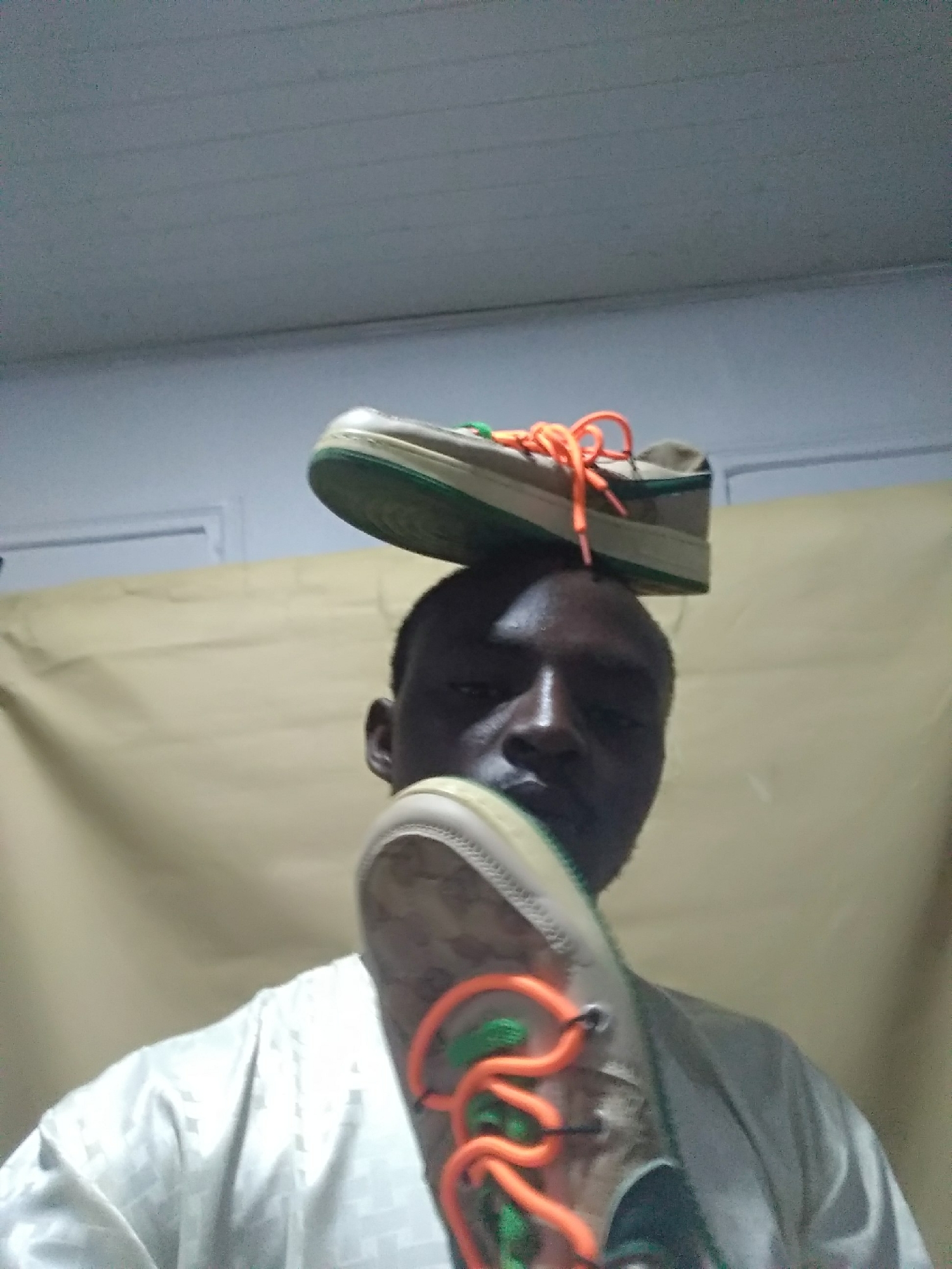 A man is pictured indoors, with one shoe balanced on his head and the other held in front of his face. The shoes are light brown with green soles and orange laces. He is wearing a white, patterned shirt. The background consists of a tan-colored sheet or curtain and a white ceiling. The location is Unknown, Unknown.