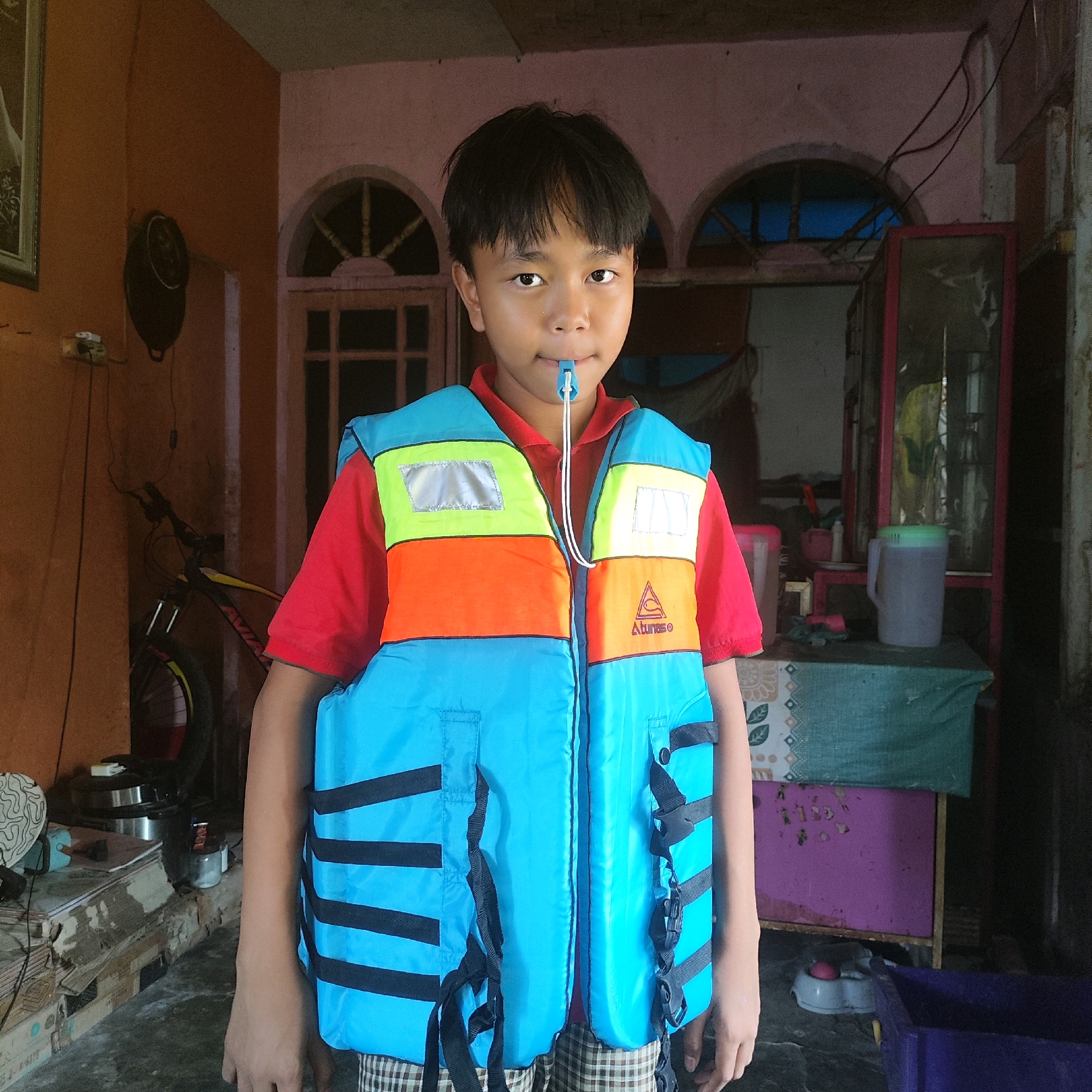 In an indoor setting that appears to be a dwelling, a young boy stands facing the camera, wearing a brightly colored life vest over a red shirt. The life vest is predominantly blue with horizontal stripes of black, and features sections of orange and lime green with reflective silver patches. A blue whistle is secured to the life vest by a white cord, and the boy has the whistle in his mouth. He is also wearing plaid shorts. The background shows an interior with doorways, windows with arched tops, and various objects including a bicycle, a framed picture on the wall, a table covered with a cloth, and large containers. The lighting suggests it is daytime, possibly with natural light coming from the open doorways or windows.