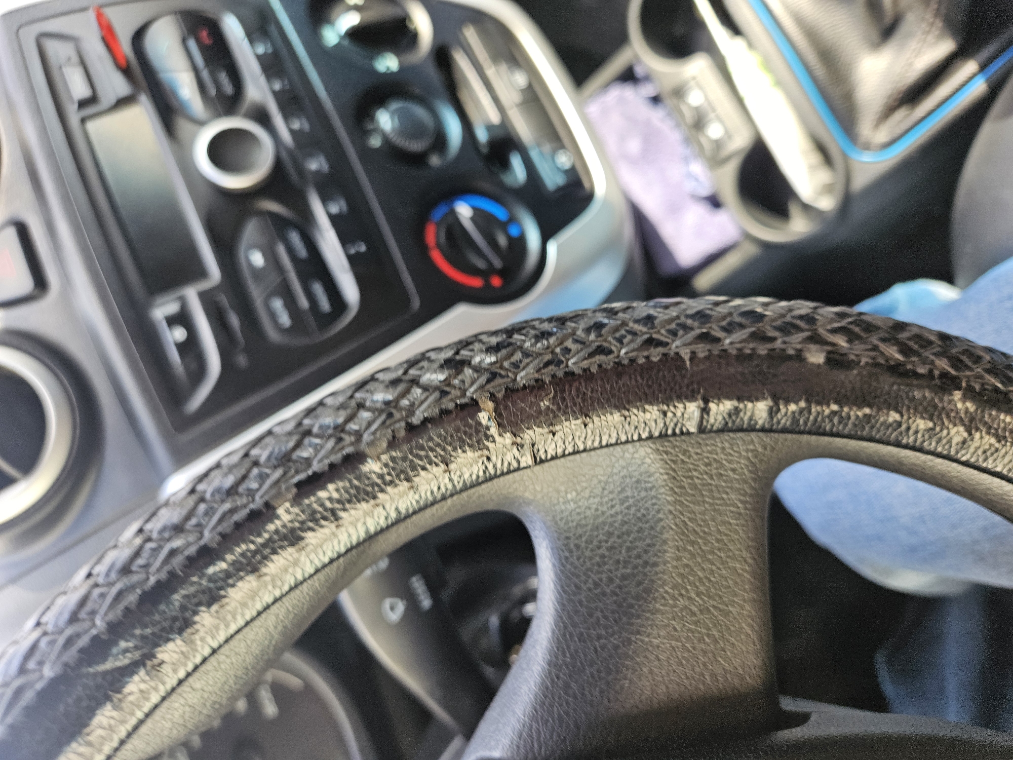 The image shows the interior of a vehicle, specifically focusing on a worn steering wheel with a peeling leather or synthetic leather cover. Behind the steering wheel, a portion of the car's dashboard is visible, including climate control knobs and other buttons. The condition of the steering wheel suggests age and wear of the vehicle. The presence of a cup holder is also noticeable. The location of the scene is not apparent from the image and cannot be determined based on the given information.