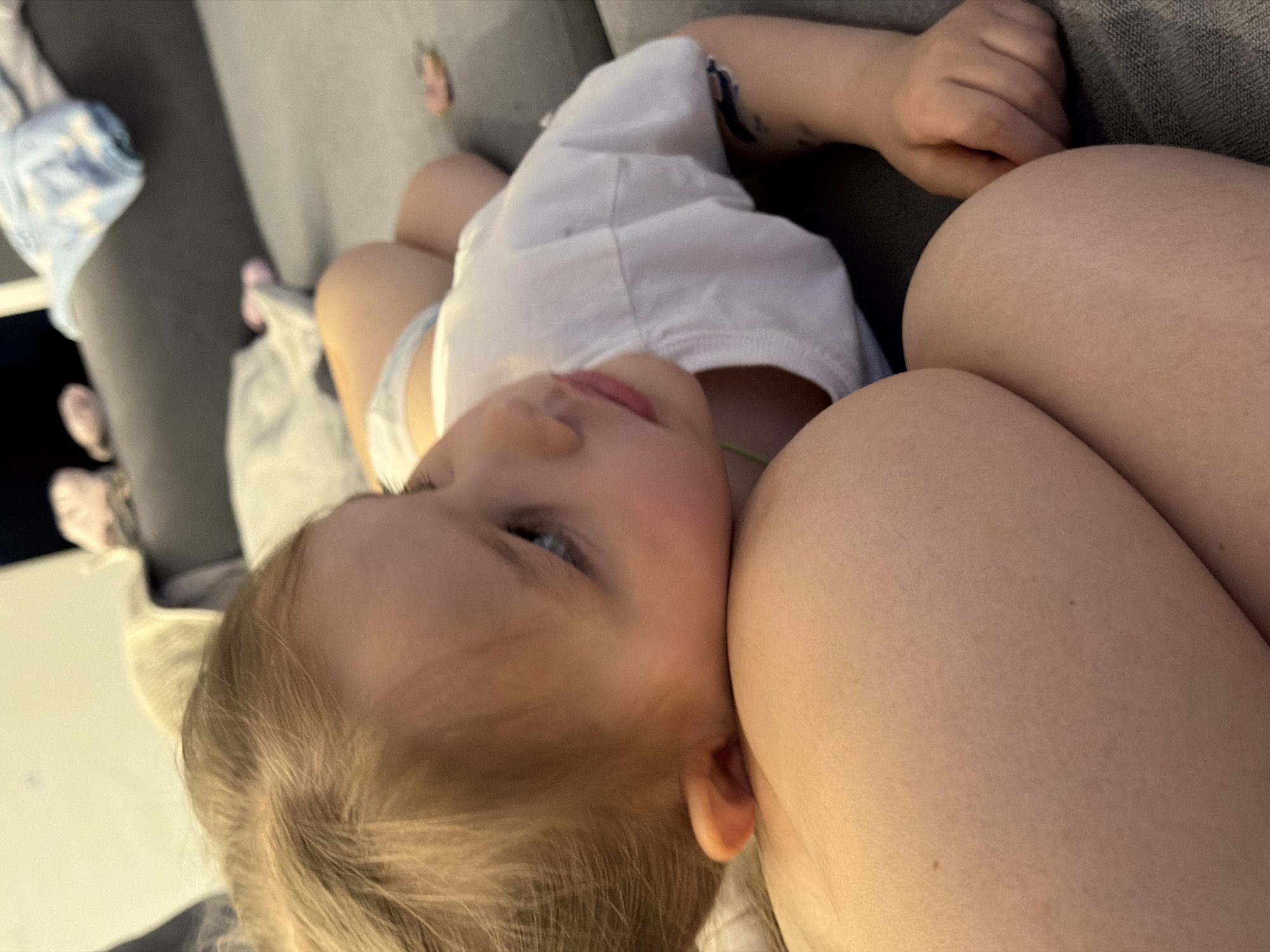 A child is lying on a person's lap. The child is fair-skinned with blonde hair and blue eyes, wearing a white shirt. The person's lap is visible, appearing to be a leg. The scene is indoors, with a gray sofa in the background. Other items like a blanket are visible on the sofa. The location, Chaikovsky, Russia, is not supported by the visual information in the image.