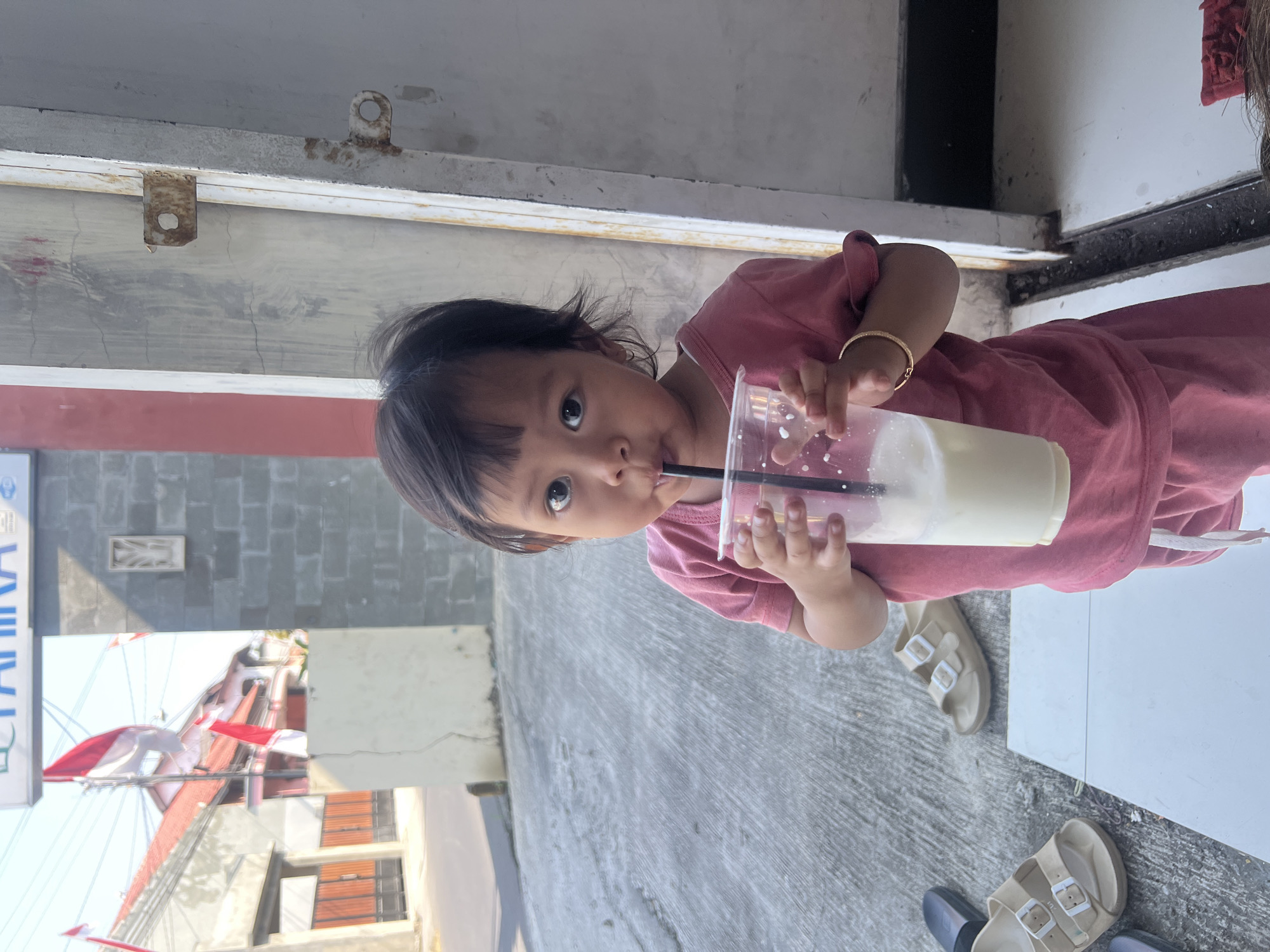 The image captures a young child drinking from a clear plastic cup with a black straw. The child is wearing a pink t-shirt and pink shorts. A gold bracelet is visible on the child's right wrist. In the background, there are houses with red-tiled roofs, some adorned with Indonesian flags. The ground is paved with concrete, and a pair of beige sandals are visible to the left of the child. To the right, a wall made of grey bricks and a white metal door frame are present. The scene appears to be an outdoor area in a residential setting, likely in Kecamatan Pati, Indonesia, given the presence of Indonesian flags and architectural elements. The lighting suggests it is daytime, with shadows indicating sunlight.
