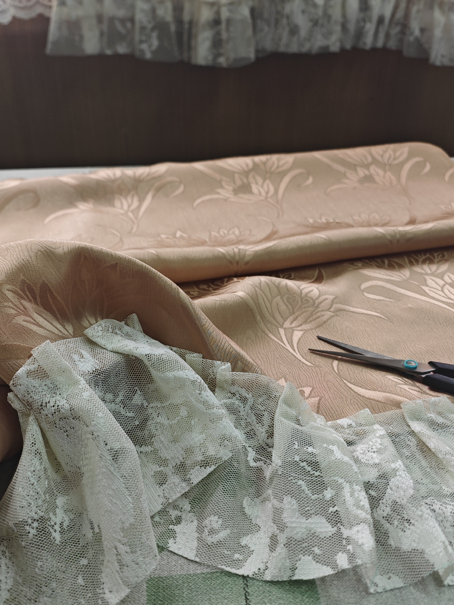The image depicts a close-up view of fabric and lace, with a pair of scissors resting on the material. The fabric appears to be a textured, light peach-colored material with a floral pattern. A piece of delicate white lace, with intricate floral designs, is draped over the fabric. The scissors are open, with black handles and a blue detail on one of the blades. The lighting suggests an indoor setting, possibly a craft room or studio. There is no discernible information in the image that would allow for the determination of a specific location within Krasnoyarsk, Russia. The scene is static and does not capture any active events or individuals.