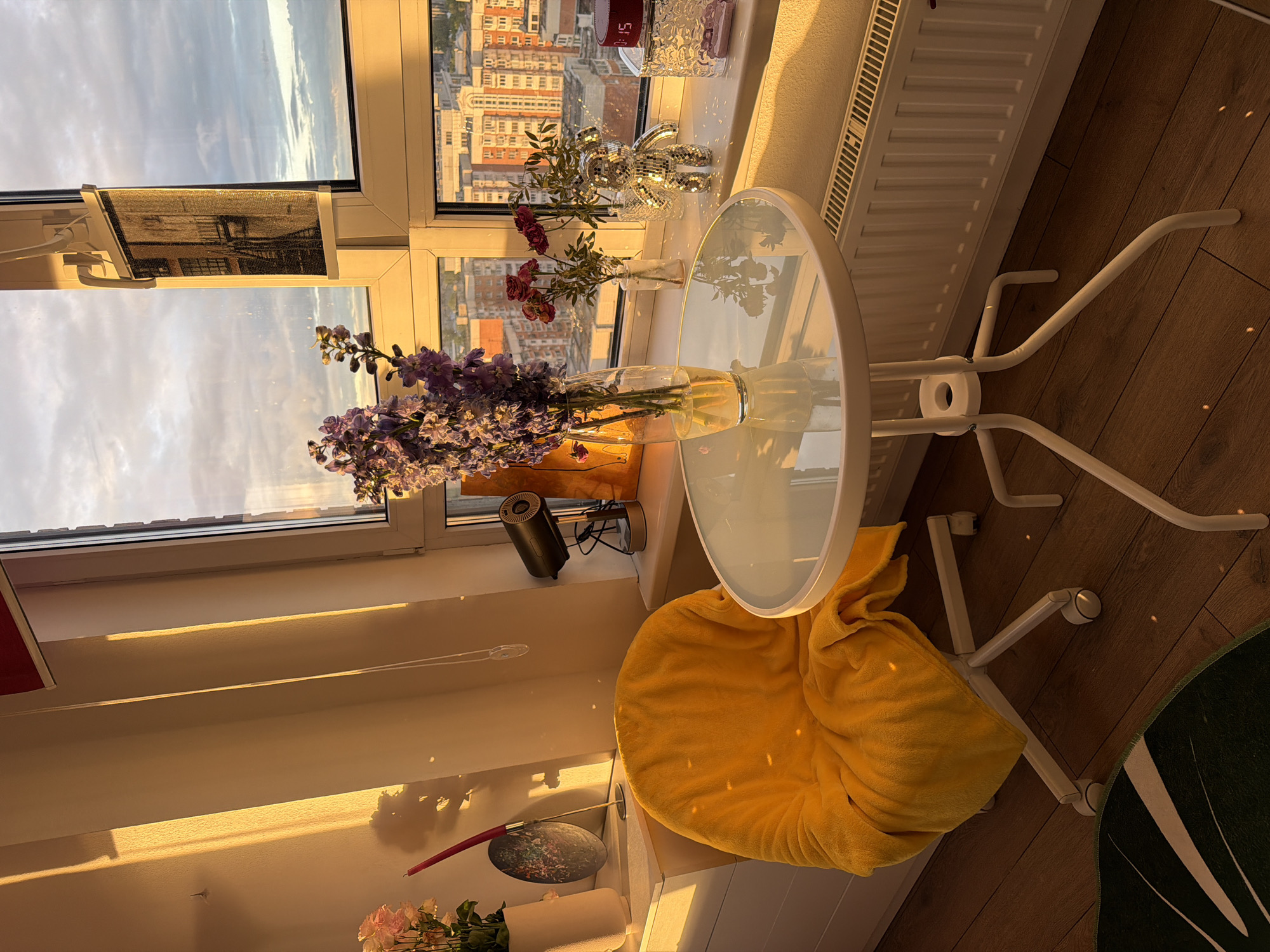The image depicts an interior scene bathed in warm sunlight, oriented vertically. Dominating the view is a window overlooking an urban landscape, with buildings visible in the distance. Several vases of flowers are arranged near the window, adding a touch of nature to the indoor setting. A small, round, glass-topped table stands next to the window, with a yellow towel or blanket draped over a chair positioned nearby. The walls are painted in a light color, and the floor appears to be wooden. The location of the city is not identifiable based on the information in this image.