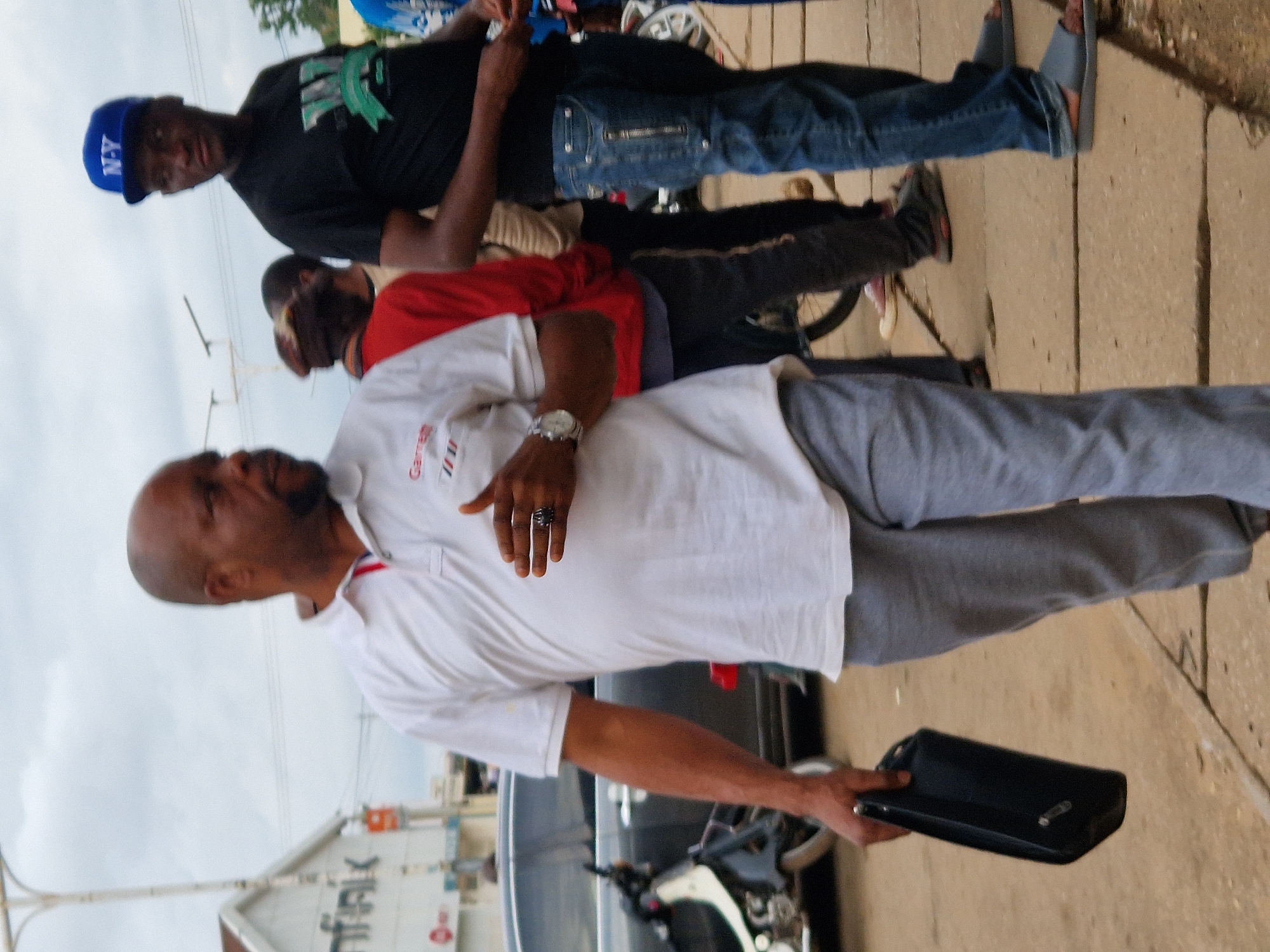 The image captures a street scene in an urban environment. In the foreground, a man with a shaved head and dark skin is walking to the left, carrying a black clutch bag in his left hand. He is wearing a white polo shirt with red and blue trim at the collar and a silver wristwatch on his left wrist. He also wears a dark ring on his right ring finger. His pants are gray. To his right, another man is walking to the right. He is wearing a black t-shirt with a green graphic, blue jeans, and gray sandals. He is also wearing a blue baseball cap with "N-Y" on it. In the background, a black car is parked, and a motorcycle is partially visible. Other people are present in the background, and buildings can be seen on the left side of the image. The sky is overcast.