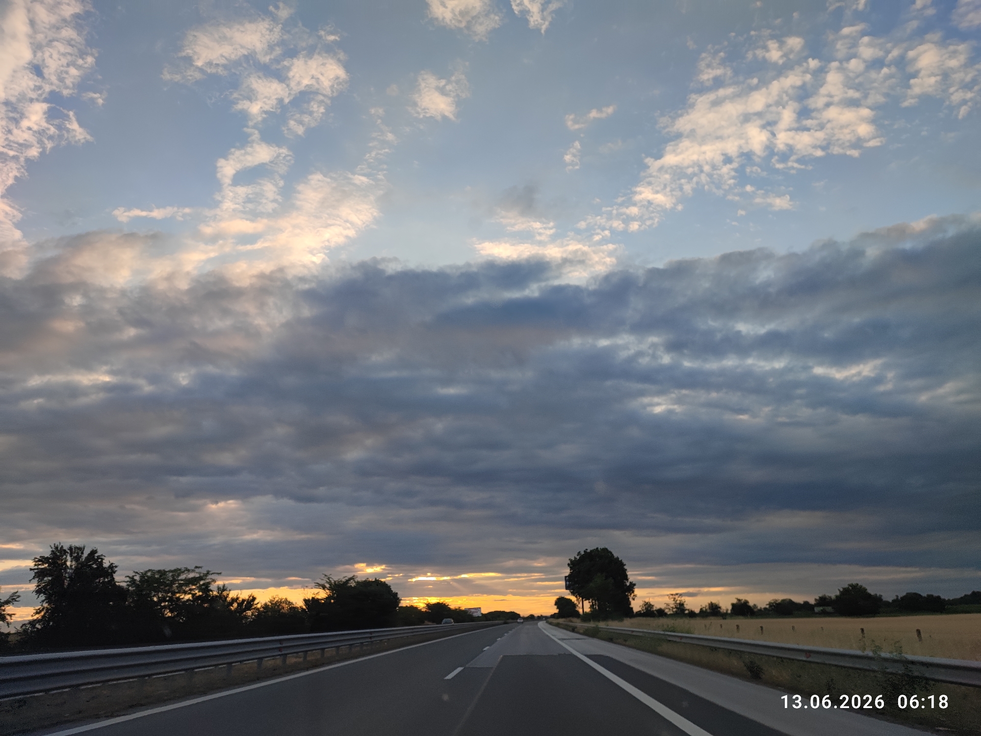The image captures a highway scene in Tsaratsovo, Bulgaria, at 06:18 on June 13, 2026. The sky is filled with dramatic clouds, a mix of dark gray storm clouds and lighter, wispy clouds tinged with the warm hues of sunrise or sunset. Sunlight breaks through the clouds in several places, casting a golden glow on the horizon. The highway itself is paved, with clear white lane markings. A metal guardrail runs along the left side of the road. On either side of the highway, there are trees and fields, with some appearing as dark silhouettes against the sky. The overall atmosphere is one of a changing sky, possibly during the early morning or late evening.