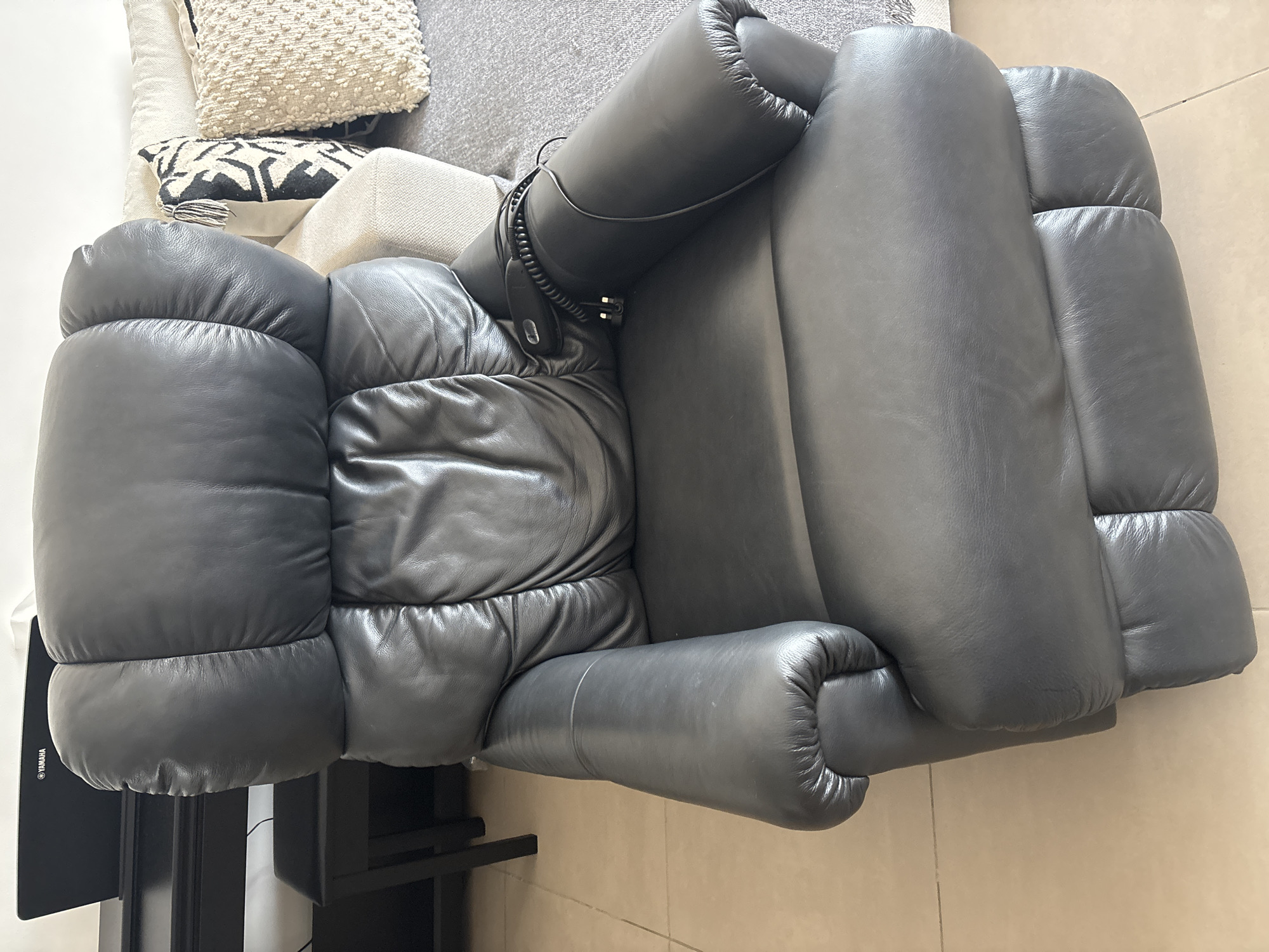 The image shows a black leather recliner chair with a wired remote control resting on its arm. The chair appears to be in a living room setting, with a sofa and decorative pillows visible in the background. A Yamaha digital piano is also partially visible in the upper left corner of the image. The floor is tiled. The scene is likely indoors in a residential setting in Dubai, United Arab Emirates.