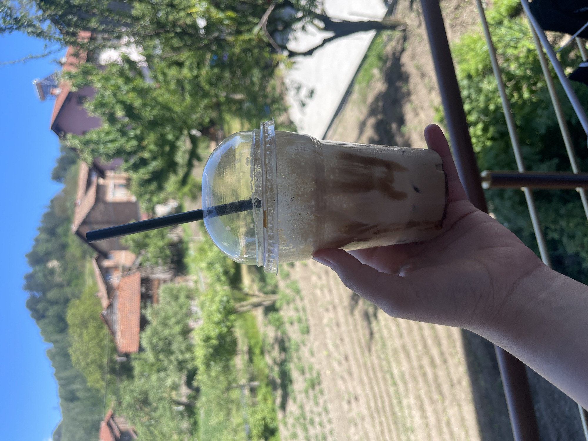 A fair-skinned hand holds a clear plastic cup of a blended coffee drink with a black straw, against a blurry backdrop showing rural Kraishte, Bulgaria. The background shows green trees and fields, residential buildings with red-tiled roofs, and a bright blue sky. A metal railing is partially visible.