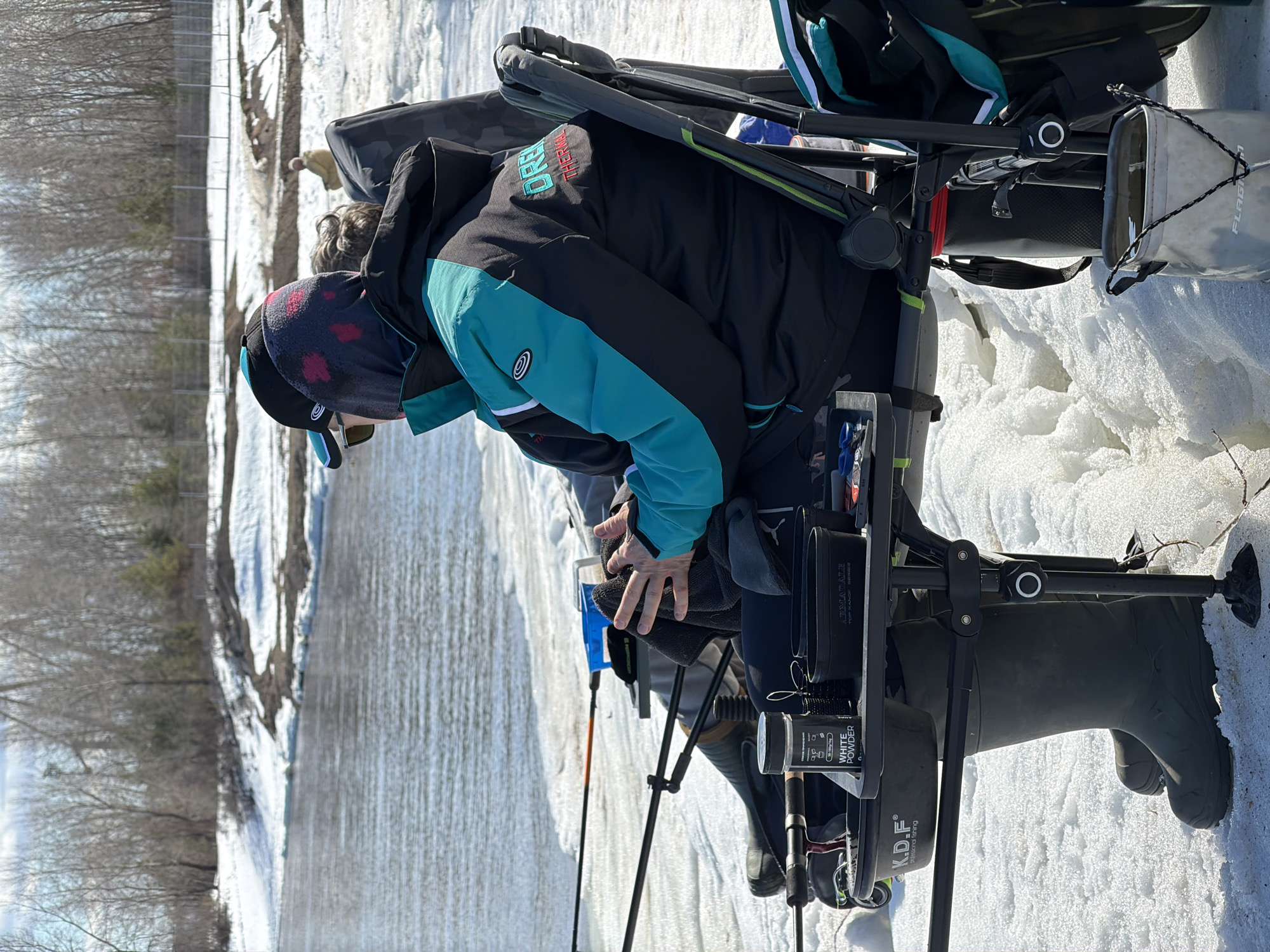 An outdoor scene depicts a person fishing on ice in a winter environment. The individual is wearing a black and teal jacket, dark headwear, and sunglasses. They are positioned on a fishing platform or chair, surrounded by gear, including fishing rods, a white powder container, and a container labeled "Armadale Top Range Series." The landscape features snow and trees. The location is uncertain, and there is no visual evidence to confirm that it is Noviy, Russia.