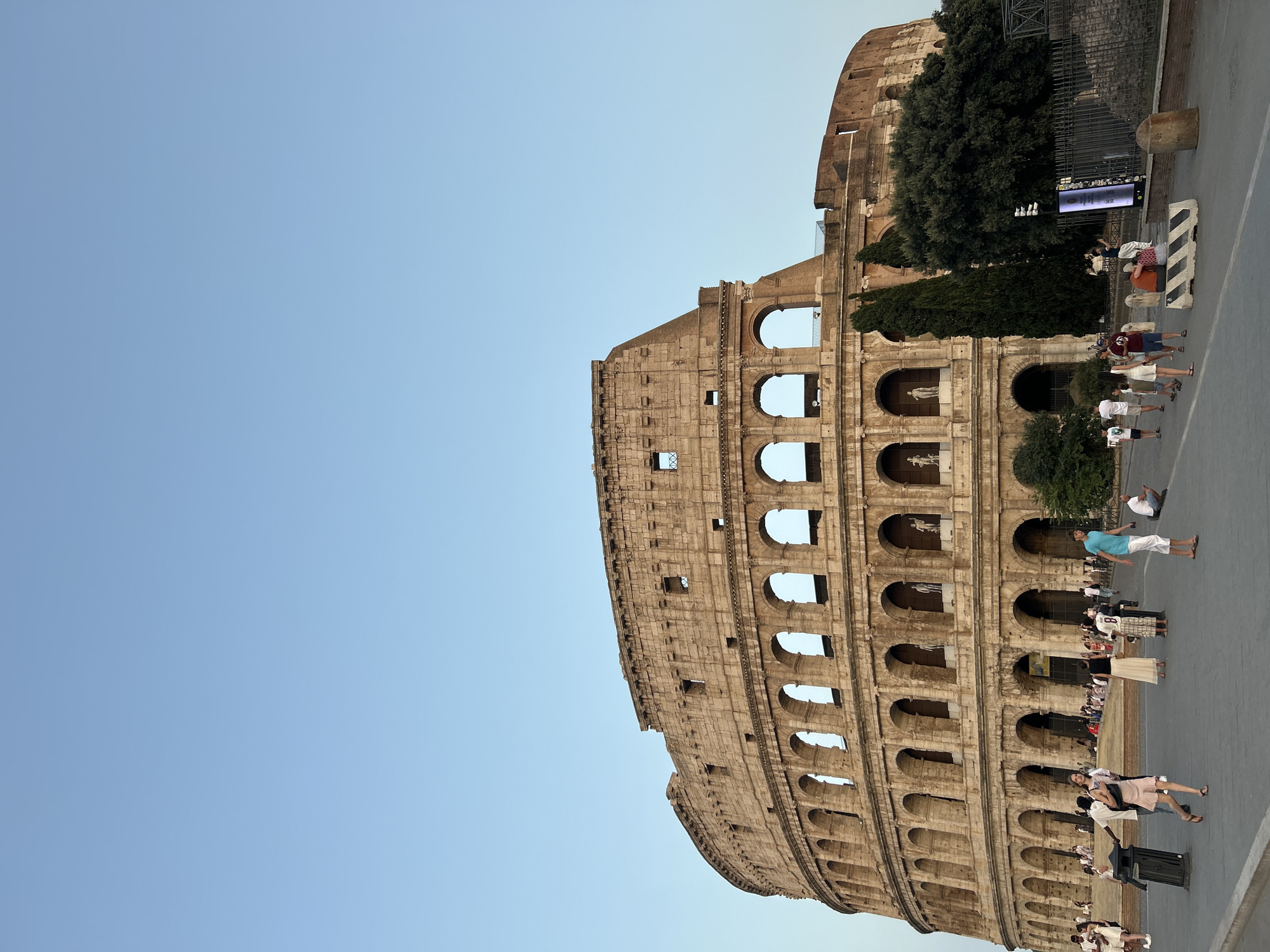 The image shows a wide, low-angle view of the Colosseum in Rome, Italy, under a clear, light blue sky. The ancient amphitheater, constructed of weathered stone, occupies the right two-thirds of the frame. Its curved, multi-tiered structure is visible, featuring numerous arches and colonnades. Several people are walking and standing on a paved area in front of the Colosseum. They are diverse in age and attire, with some appearing to be tourists. To the right of the Colosseum, there are trees and a modern digital display board. The lighting suggests it is either early morning or late afternoon.