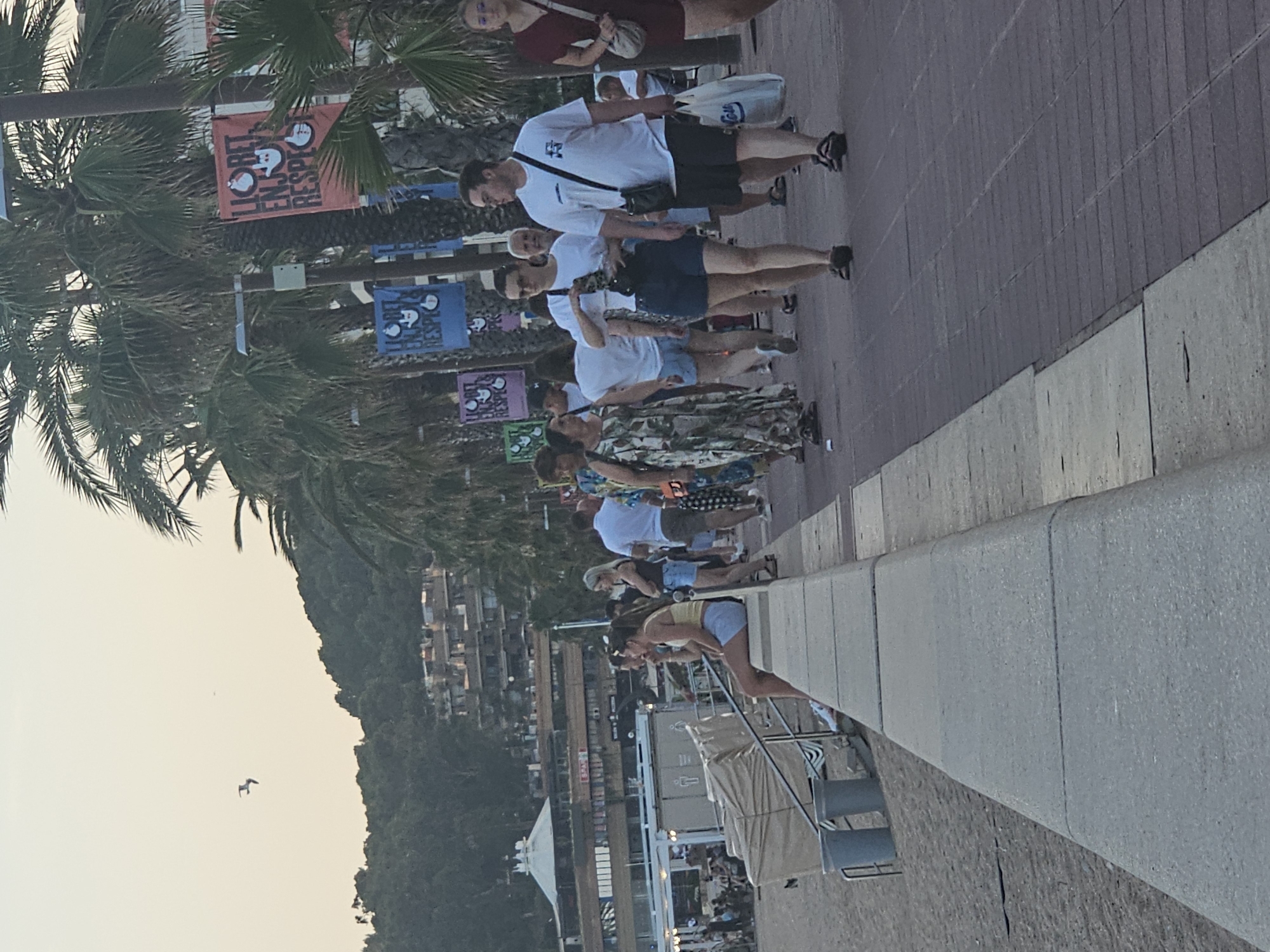 This image captures a scene along a promenade in Lloret de Mar, Spain. A group of people are walking on a paved walkway beside a beach. To the left of the walkway, there is a concrete wall bordering the sandy beach. In the background, there are buildings nestled on a hillside and palm trees line the promenade. Several banners are hanging from lampposts, featuring graphics and text that appear to be related to the location. Some individuals are casually dressed, while others are carrying shopping bags. The overall atmosphere suggests a relaxed, possibly tourist-oriented setting, during daylight hours.