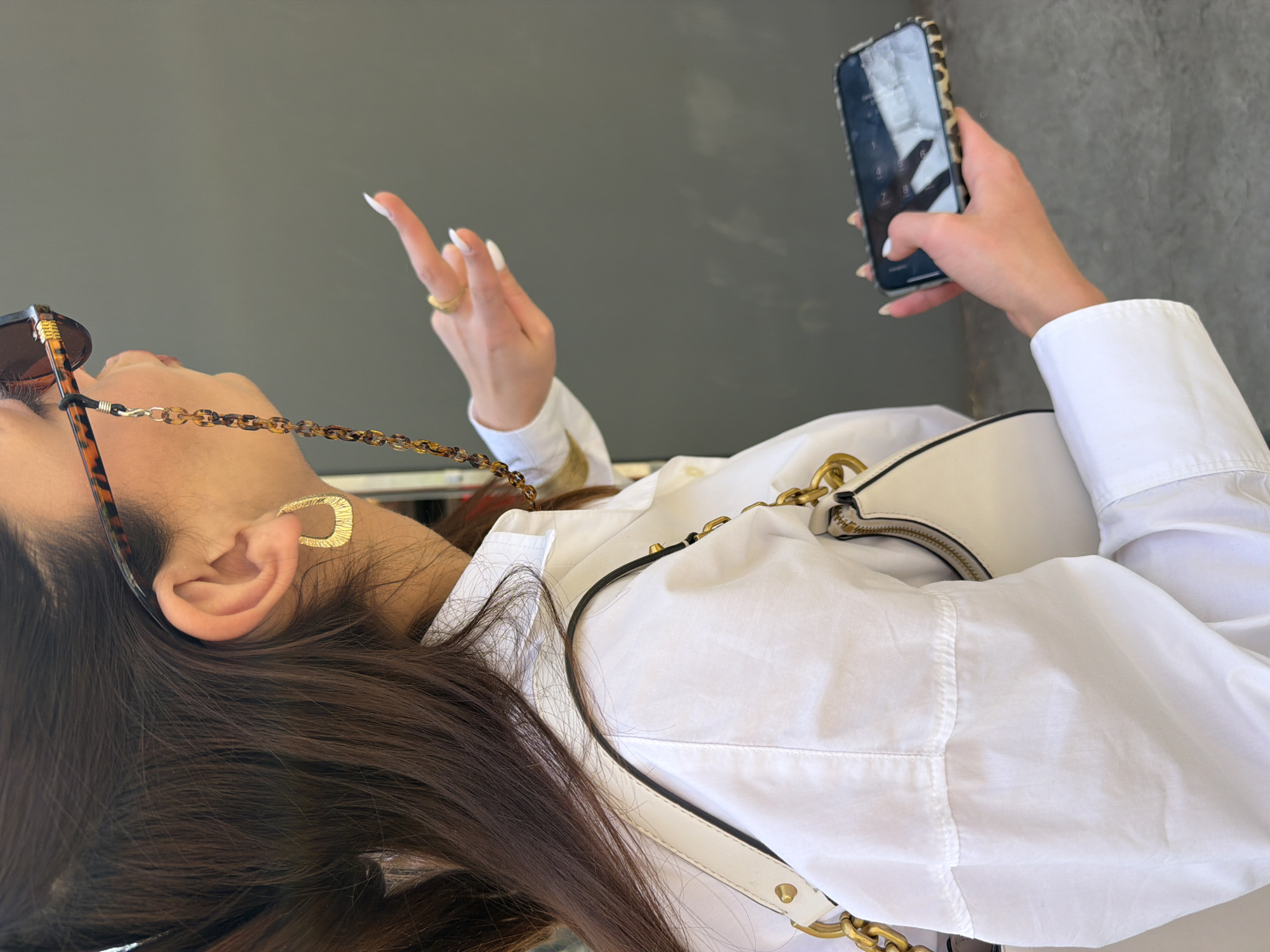 The image captures a close-up, rotated view of a person engaged with their phone against a plain, dark gray background, likely in Yerevan, Armenia.
The subject has long, dark brown hair and is wearing a crisp white button-down shirt. A pair of tortoiseshell-patterned sunglasses hangs around their neck, connected by a matching chain. A prominent gold, U-shaped earring adorns their left ear. On their left hand, a golden ring is visible on the ring finger, and their fingernails are long and painted white. This hand is raised, fingers slightly splayed, possibly in a gesture of speech or direction.
In their right hand, the person holds a smartphone encased in a tortoiseshell or leopard print case. A finger is positioned on the screen, which shows a faint reflection of a keypad or interface, indicating they are likely interacting with the device, perhaps unlocking it or making a selection. A white handbag with a distinctive gold chain strap and a gold zipper is slung over their shoulder.
The setting appears to be a well-lit area during the daytime, though no direct sunlight is evident. The background is simple and devoid of specific environmental cues beyond its uniform dark gray color. No visible text is discernible in the image.