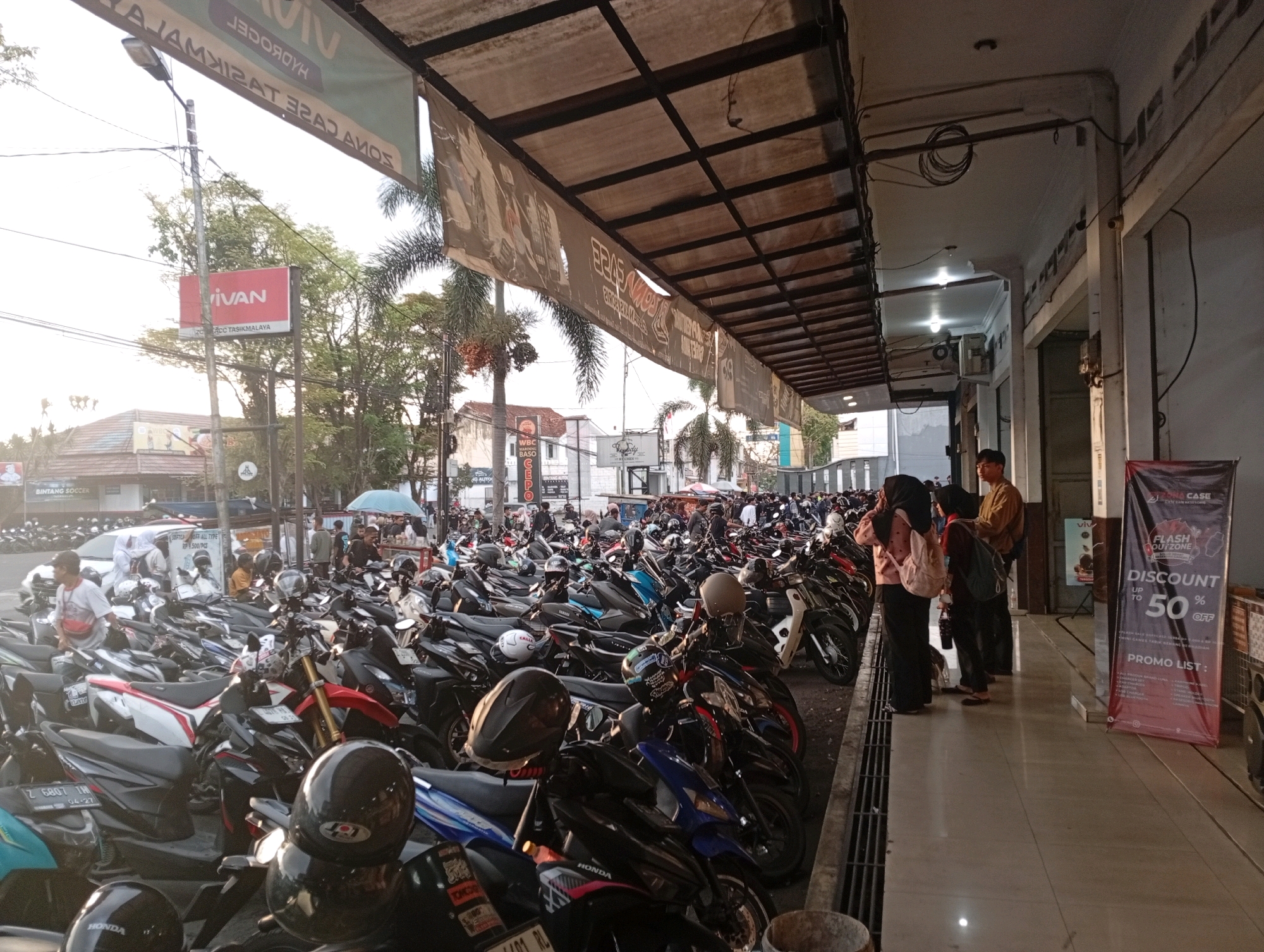 The image depicts a busy street scene in Tasikmalaya, Indonesia, characterized by a large number of parked motorcycles. The foreground and midground are dominated by a dense collection of scooters and motorcycles, suggesting a parking area or a busy commercial street. Several people are visible, some standing or walking amongst the vehicles, while others are gathered near a storefront. One group of individuals, including two women and a man, are standing on a sidewalk, possibly interacting or waiting. To the right of the image, a large promotional banner displays "FLASH OUTZONE" and "DISCOUNT UP TO 50% OFF," indicating a commercial establishment or event. The environment appears to be outdoors, with a covered walkway or awning overhead supported by metal structures. Buildings line the street, and trees are visible in the background, suggesting an urban setting. The lighting indicates it is daytime, possibly late afternoon given the angle of the sun. The overall impression is one of a bustling commercial or service area, typical of Indonesian cities, with a high density of two-wheeled transportation.