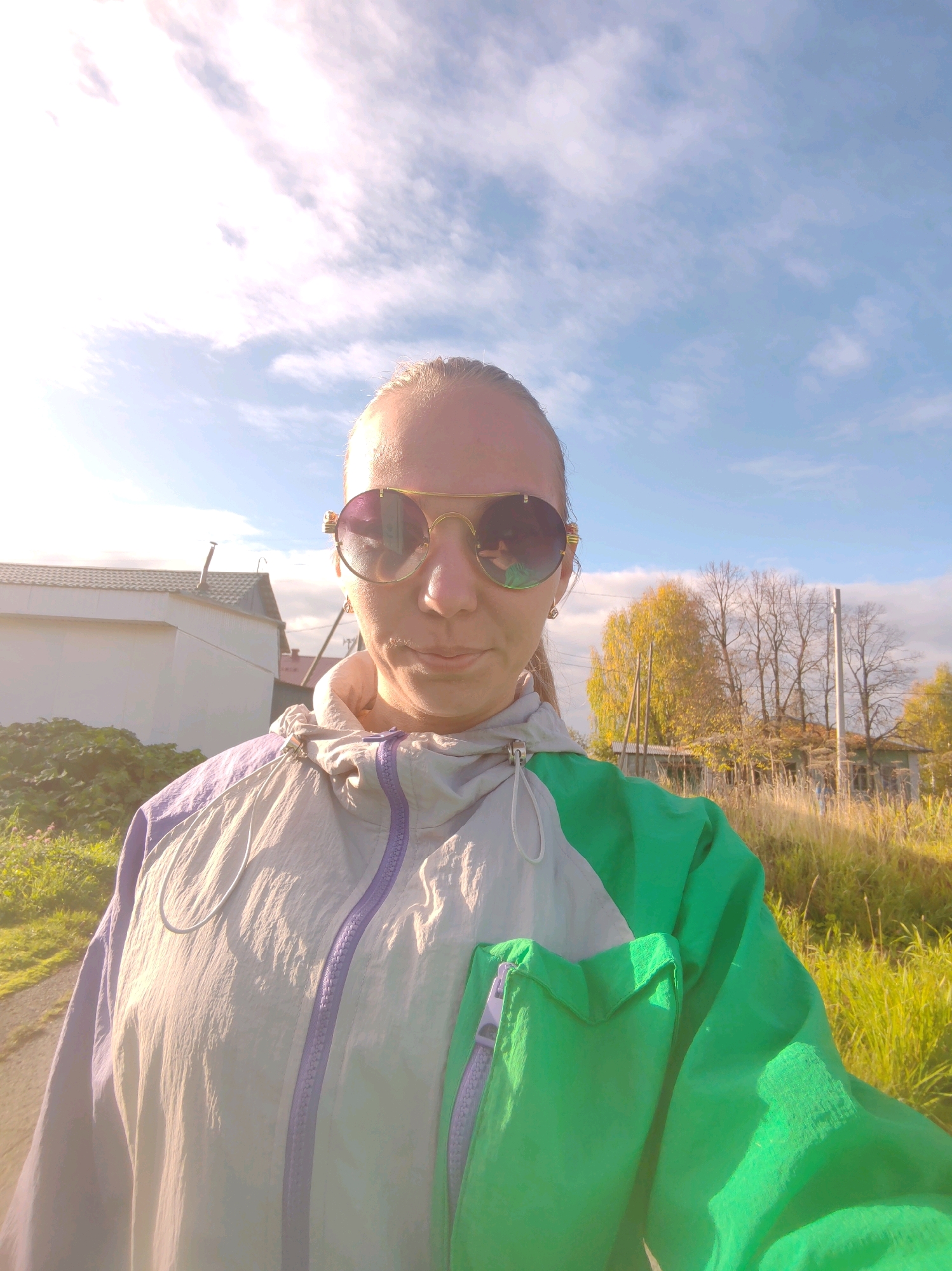 A person is taking a selfie outdoors on a sunny day, with a bright blue sky and scattered clouds. They are wearing large, round sunglasses with a gold frame and a multi-colored jacket. The jacket is primarily light gray with lavender and green accents, featuring a zippered front and pockets. In the background, there is a white building with a dark roof and a small chimney. To the right, there are trees with yellowing leaves, indicating autumn, and a grassy field. The general environment suggests a rural or suburban setting, consistent with the city of Chukhloma, Russia. The lighting indicates it is daytime.