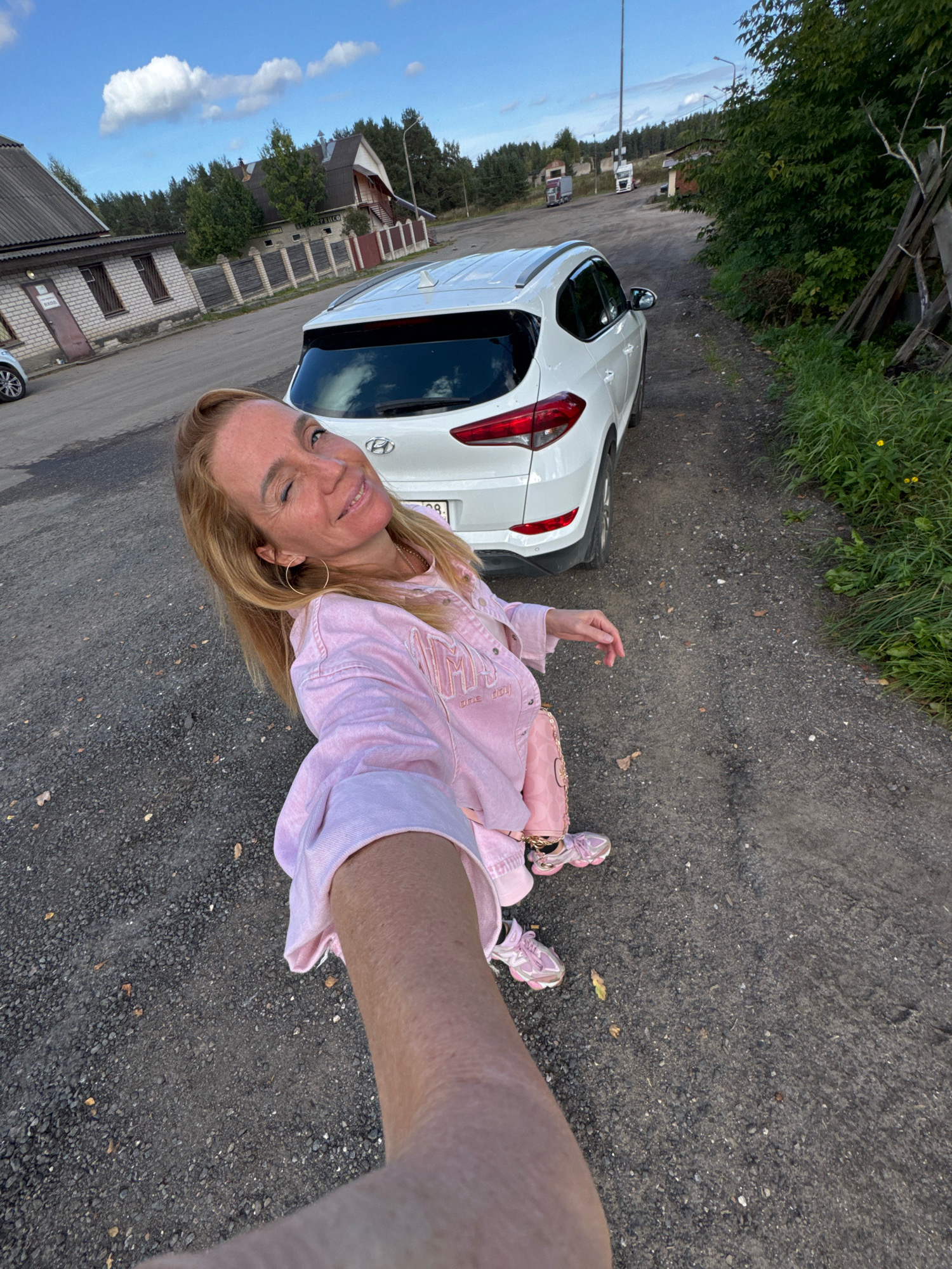 In Novyi Berezai, Russia, a blonde woman with fair skin and freckles takes a selfie outdoors. She is wearing a light pink tracksuit and pink sneakers. Her left arm is extended towards the camera, and she is smiling and winking. Behind her, a white Hyundai SUV is parked on a gravel surface. The background features a building with a grey roof and brick walls, a wooden fence, and trees. The sky is blue with scattered white clouds. A tall pole with lights is visible in the distance.