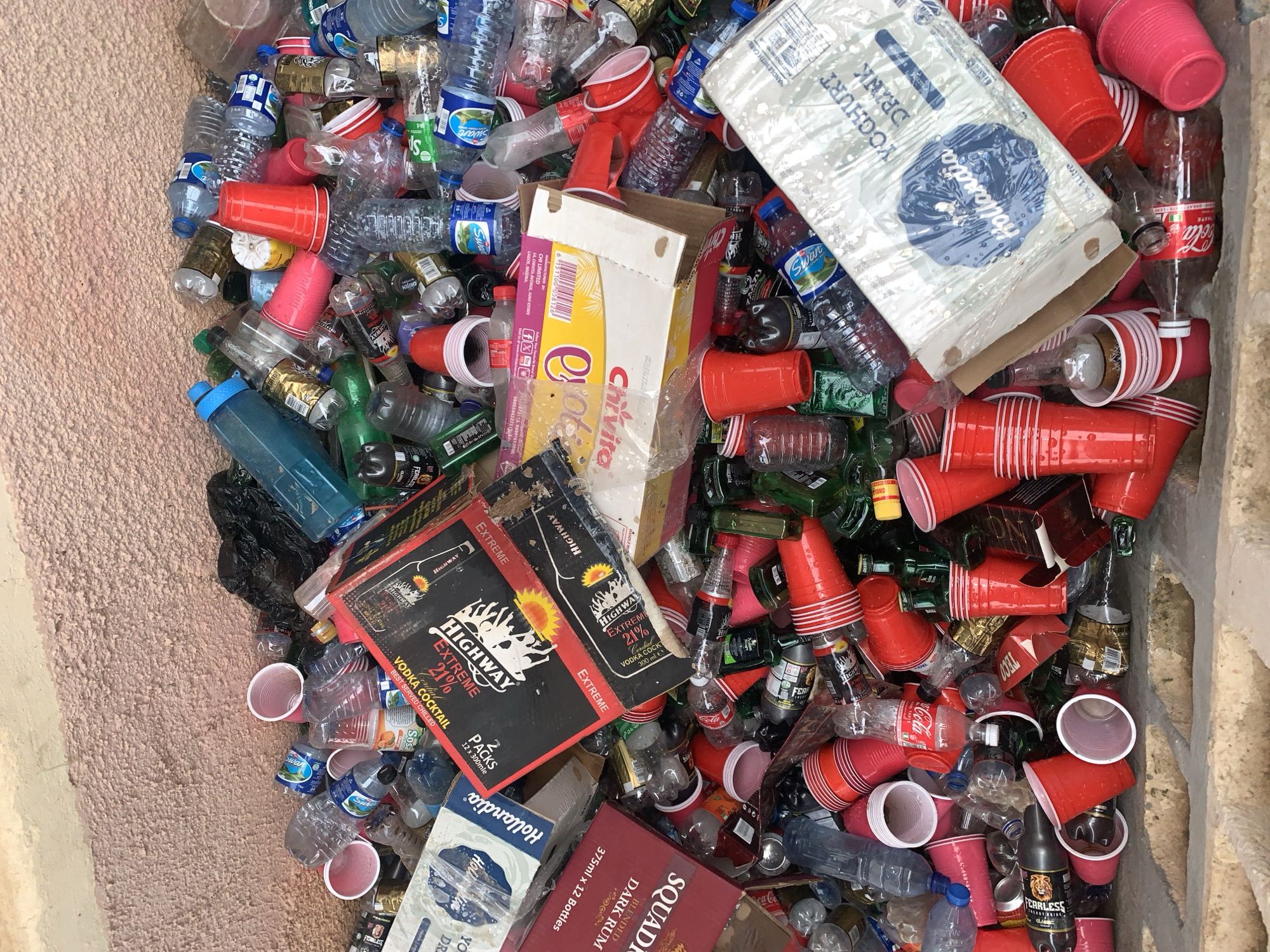 A top-down shot depicts a refuse pit, possibly within the vicinity of a bar or social gathering area, located in or near New Stadium, Nigeria, containing a multitude of discarded items, predominately single-use plastic and paper waste. Red disposable plastic cups are abundant. Empty plastic water bottles branded "Swan" and "Fearless" beer bottles are visible, along with assorted plastic and glass beverage containers. Various discarded packaging boxes are scattered throughout the pile. Packaging from "Hollandia Yoghurt Drink", "Chivita", "Highway Extreme Vodka Cocktail", and "Squadra Dark Rum" are distinguishable among the waste. The pit is likely surrounded by walls of tan concrete. No people are visible.