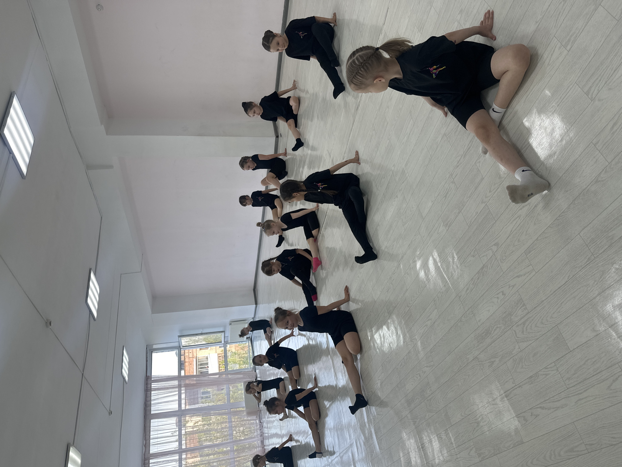 A group of approximately 13 children are practicing a dance routine in a studio. They are wearing black dance attire, some with socks. The studio has light-colored wooden floors and white walls with a row of windows along one side, revealing a view of the outside - including buildings and trees. Linear LED lights are attached to the wall near the ceiling. The location may be Votkinsk, Russia. The children are bending forward, with one hand on the floor and the other extended. They are arranged in rows.
