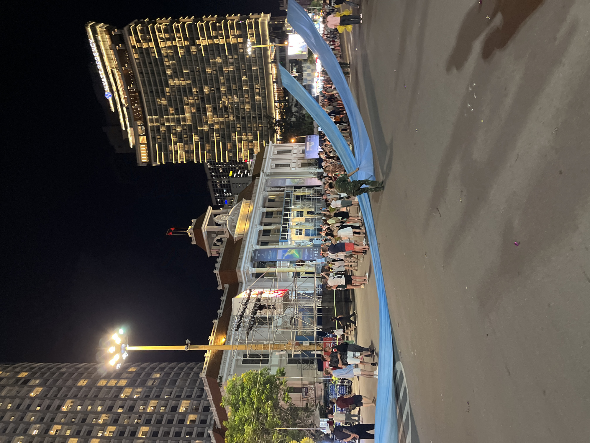 In Nha Trang, Vietnam, at night, a large, light blue fabric is being unfurled across a street in front of a grand, white building with classical architectural elements and a red-tiled roof. The building is illuminated, and scaffolding is visible on its facade, suggesting ongoing construction or an event setup. On the right side of the frame, a modern, curved skyscraper with numerous illuminated windows, identified by a "HYATT REGENCY" sign, towers over the scene. A military-dressed individual is visible holding one end of the blue fabric. A crowd of people, dressed in casual attire, stand behind the fabric, observing the unfolding event. Streetlights cast yellow light on the asphalt road, which shows scattered debris. The sky is dark, indicating nighttime.